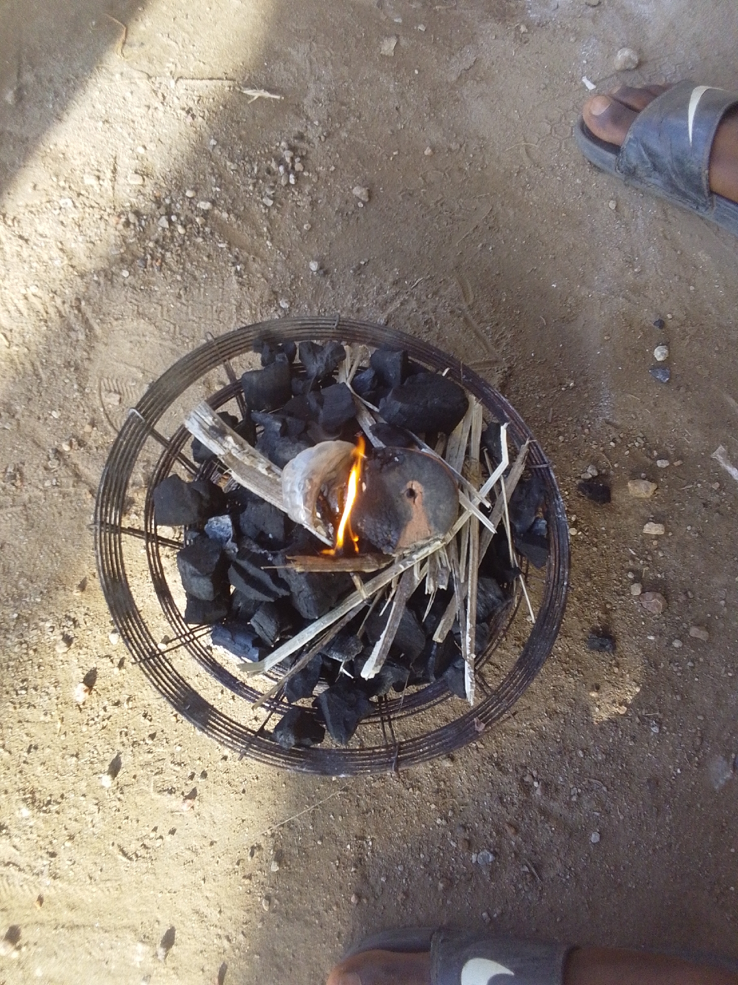 This overhead shot depicts an outdoor scene in Keffi, Nigeria, focusing on a circular metal charcoal burner placed directly on light brown, sandy ground mixed with small pebbles and debris. The burner, constructed from concentric rings of metal wire or rebar, contains numerous irregularly shaped pieces of black charcoal. A small fire is active within the burner, characterized by an orange-yellow flame emanating from a small, dark, possibly metallic or ceramic object placed amidst the charcoal and thin, light-colored wooden kindling splinters.
To the upper right of the burner, the bare foot and lower leg of an individual, wearing a dark grey sandal with a white Nike swoosh logo, are partially visible. Another leg, also wearing a similar dark grey Nike sandal, is partially visible at the bottom center-right of the frame. The ground shows subtle indentations and minor scattered detritus. Strong natural light from an unseen source casts a distinct shadow diagonally across the upper left portion of the image, indicating daylight conditions.
