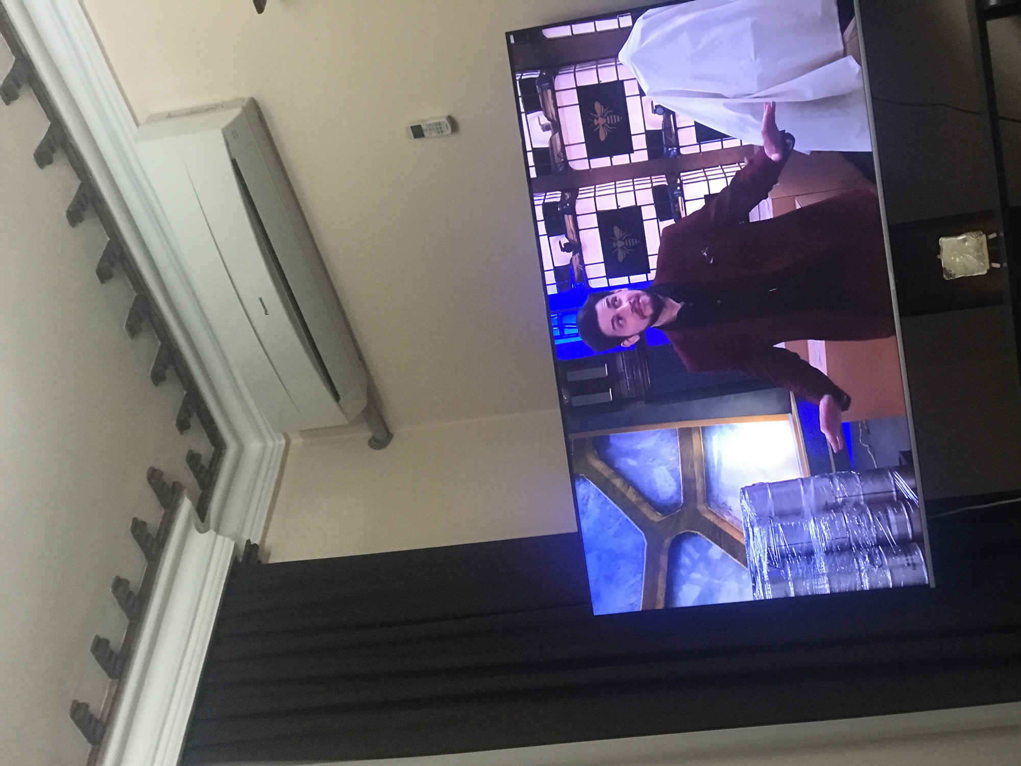 A TV is mounted on a wall with a black curtain below it. On the screen, a man with dark hair and a beard, wearing a maroon jacket, is gesturing with both hands. The background of the TV screen shows shelves with decorations featuring a bee emblem. Above the TV, there is a white air conditioning unit attached to the wall. To the left, there is decorative molding along the ceiling. The scene gives no information to indicate it is in Yerevan, Armenia.