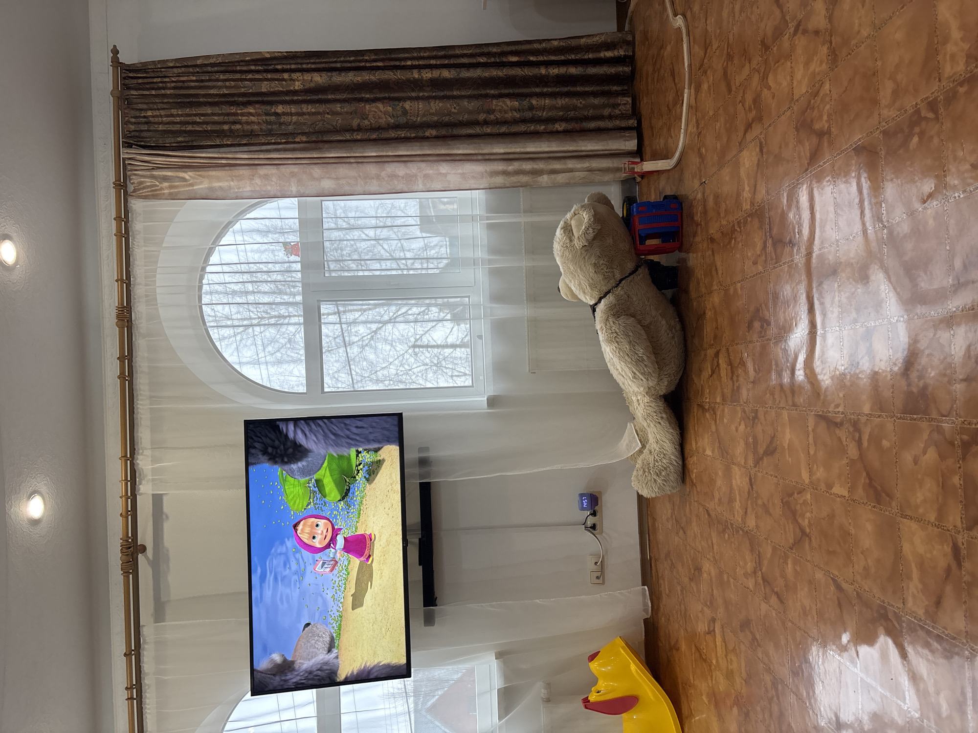 A room interior features a television showing an animated program with a character resembling Masha from "Masha and the Bear". A large teddy bear stands beside the window. The room has tiled flooring, white walls, and a window with a semi-circular top. Brown curtains adorn the window. Toys, including a yellow slide, are scattered around the room. There are no people visible. The location appears to be a domestic setting, possibly in Ferma, Russia.