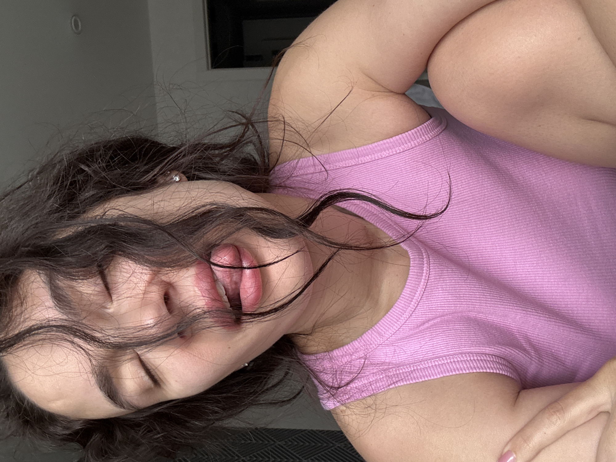 A woman with dark hair wearing a pink tank top is lying down. Her eyes are closed, and her lips are slightly parted. She is indoors in a room with a window visible in the background. The location is unknown, and there is no evidence to suggest it is Chaikovsky, Russia.