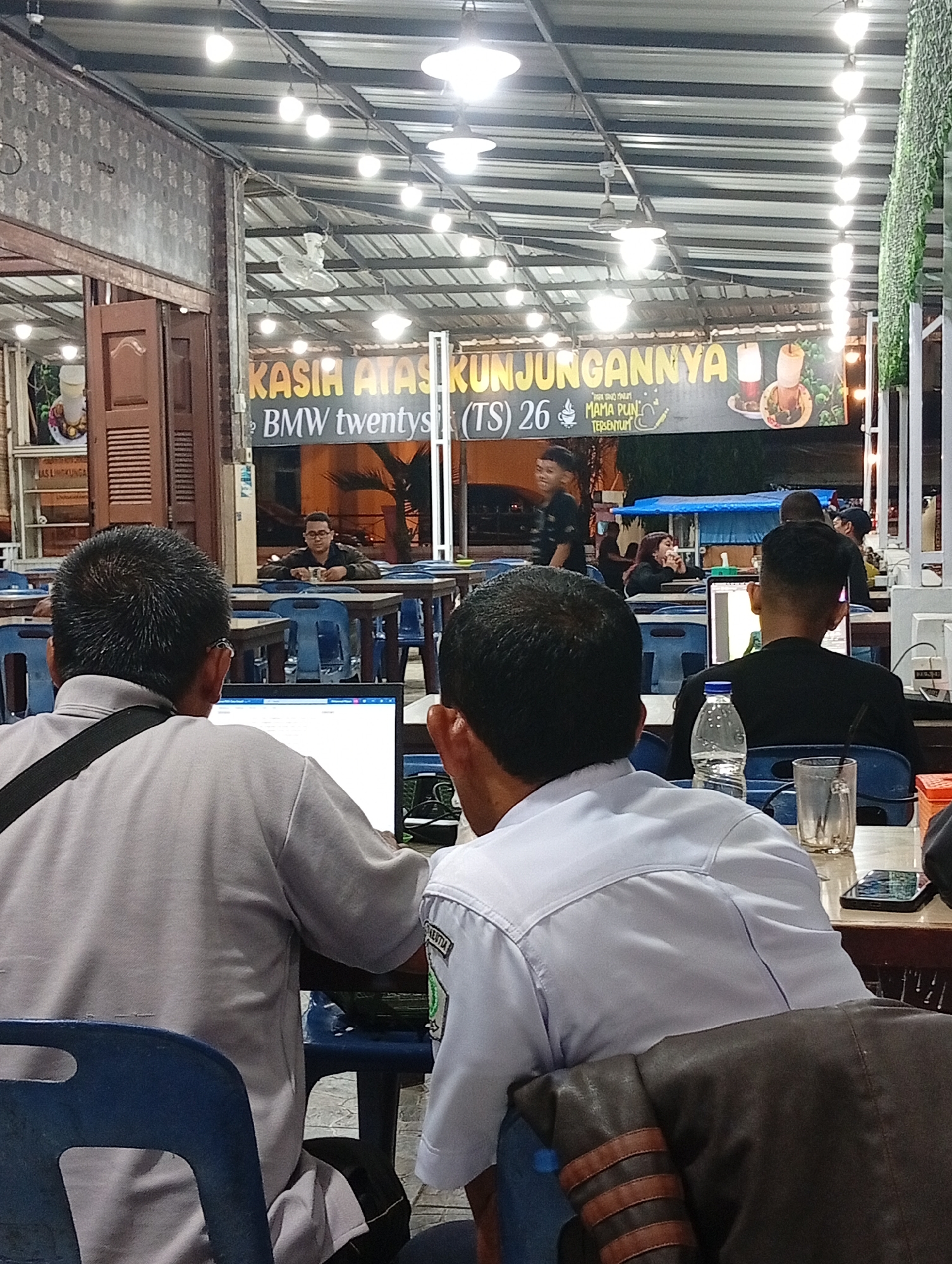 This image captures a nighttime scene inside what appears to be a casual restaurant or cafe in Lhokseumawe, Indonesia. The venue features a high ceiling with exposed corrugated metal and numerous bare light bulbs providing bright illumination.
In the foreground, two men are seated at a wooden table, viewed from behind. The man on the left, wearing a light-colored polo shirt and a black messenger bag, is engrossed in a laptop, its screen visible displaying a Windows interface. Beside him, the man on the right, in a light blue collared shirt, is also focused on a laptop screen. On their table, a clear plastic water bottle, a glass, and a smartphone are visible. A brown jacket with three horizontal light brown stripes is draped over the back of the right man's blue plastic chair. Both men appear to be working or engaged with their laptops.
In the mid-ground, other patrons are scattered around. On the left, a man in glasses sits alone at a table, holding a cup. Further back, several other individuals are present, some dining, others interacting. A person in a black t-shirt is seen walking past. The scene suggests a relaxed atmosphere where people can dine and work.
A large banner hangs prominently in the background. The visible text on the banner reads: "KASIH ATAS KUNJUNGANNYA" (Thanks for your visit). Below this, it continues with "BMW twentysi(TS) 26" and "MAMA PUN TERSE(NYUM)", with the end of "TERSENYUM" (smiles) cut off by a structural pole. An image depicting two drinks is also visible on the right side of the banner.