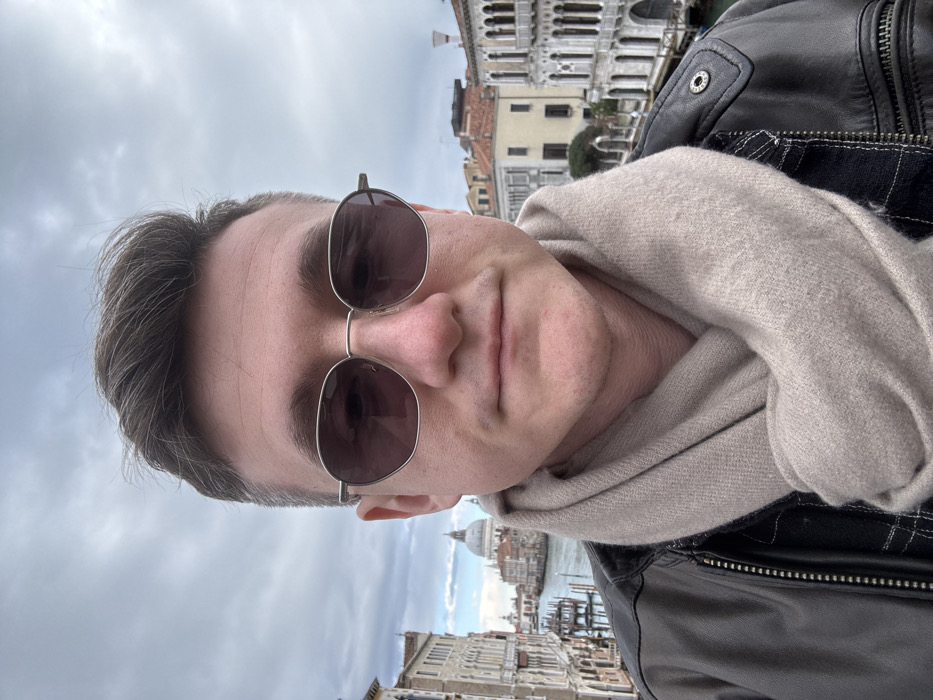The image presents a headshot-style portrait of a light-skinned male with short, dark hair, wearing dark-tinted hexagonal sunglasses, a beige knitted scarf, and a dark leather jacket over a dark plaid shirt. He is positioned centrally, facing forward with a neutral expression.
Behind him, the city of Venice, Italy, is visible under an overcast sky. Over his left shoulder, the Grand Canal stretches into the distance, leading to the prominent dome and a portion of the facade of the Basilica di Santa Maria della Salute, located in the Dorsoduro district. Historic Venetian buildings with multiple stories and arched windows line the canal, and dark wooden mooring posts are visible in the water. Over his right shoulder, further Venetian architecture is present, featuring ornate multi-story buildings alongside a waterway. The perspective strongly suggests the photograph was taken from the Ponte dell'Accademia, overlooking the Grand Canal towards the Basilica di Santa Maria della Salute.