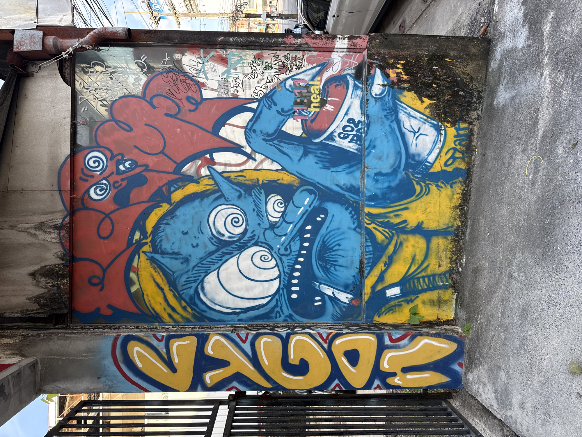 A close-up, eye-level, outdoor, natural light, full shot depicts a vibrantly painted wall in Phuket City Municipality, Thailand. The graffiti art dominates the frame, featuring a blue, cartoonish monster with large, spiraling eyes, sharp teeth, and a cigarette hanging from its mouth. The monster is depicted with a large red cloud-like shape behind its head, and its blue hand holds a spray can. The wall itself is weathered and stained, with some visible grime and moss. To the left of the graffiti, a black metal fence with vertical bars is visible. The ground in front of the wall is a concrete pavement, with some small weeds and fallen leaves. In the background, glimpses of a street, electrical wires, and a white car can be seen, suggesting an urban environment. The overall scene captures a section of an urban landscape adorned with striking street art.