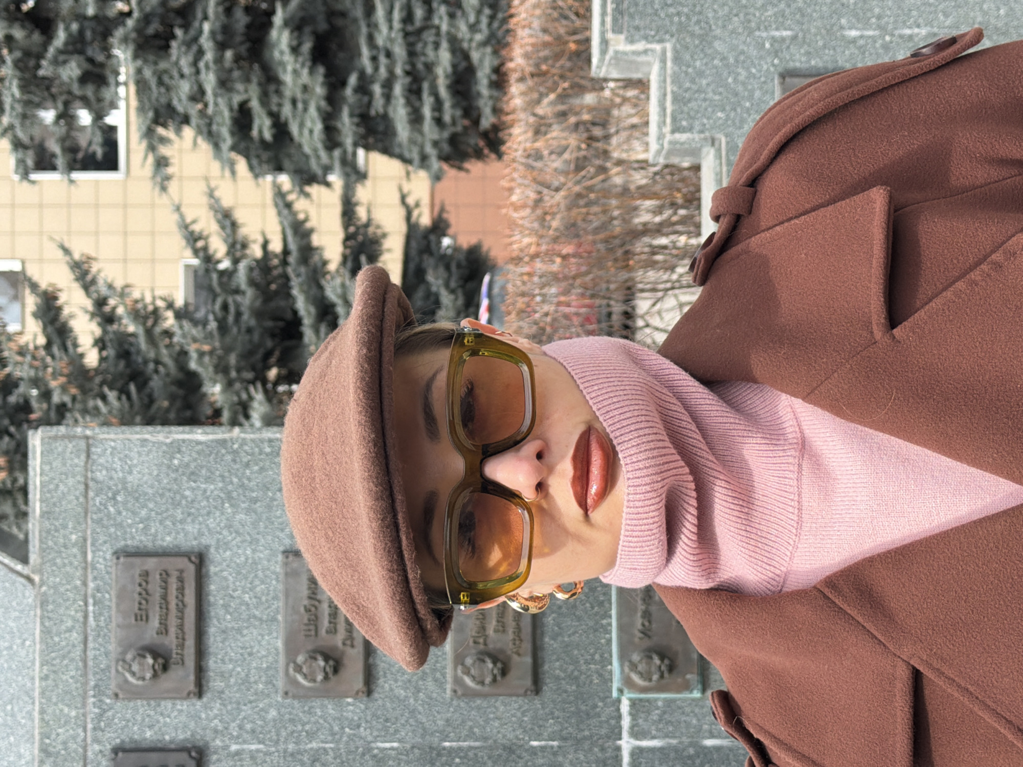 A portrait-style photograph, originally oriented vertically but depicting a horizontal view, features a person, likely female, posing outdoors in Krasnoyarsk, Russia. The individual is shown from the chest up, looking directly at the camera. She is wearing a brown flat cap, large square-framed sunglasses with yellowish-brown lenses, a light pink ribbed turtleneck sweater, and a brown wool coat. A prominent gold hoop earring adorns her right ear, and her lips are coated with a glossy, brownish-red lipstick.
The background on the left side of the image features a light yellow building with visible windows, partially obscured by dense evergreen trees. To the right, there are sparser trees with bare branches and a grey stone structure, which appears to be a monument or memorial wall.
Set into this grey stone structure are several rectangular bronze plaques with Cyrillic inscriptions. One plaque clearly displays the text "Егоров Владимир Владимирович" (Egorov Vladimir Vladimirovich). Another plaque to its right is partially visible with the beginning of a name, "Шобуни...". Additional plaques are visible below these, but their text is less clear due to focus and cropping. These plaques suggest the setting is a memorial site.
The lighting is soft and even, characteristic of an overcast day or diffused sunlight, and no harsh shadows are visible. The clothing worn by the subject indicates cool weather, aligning with an autumn or winter season in the region. The overall image blends a fashionable personal portrait with a historical or commemorative backdrop.