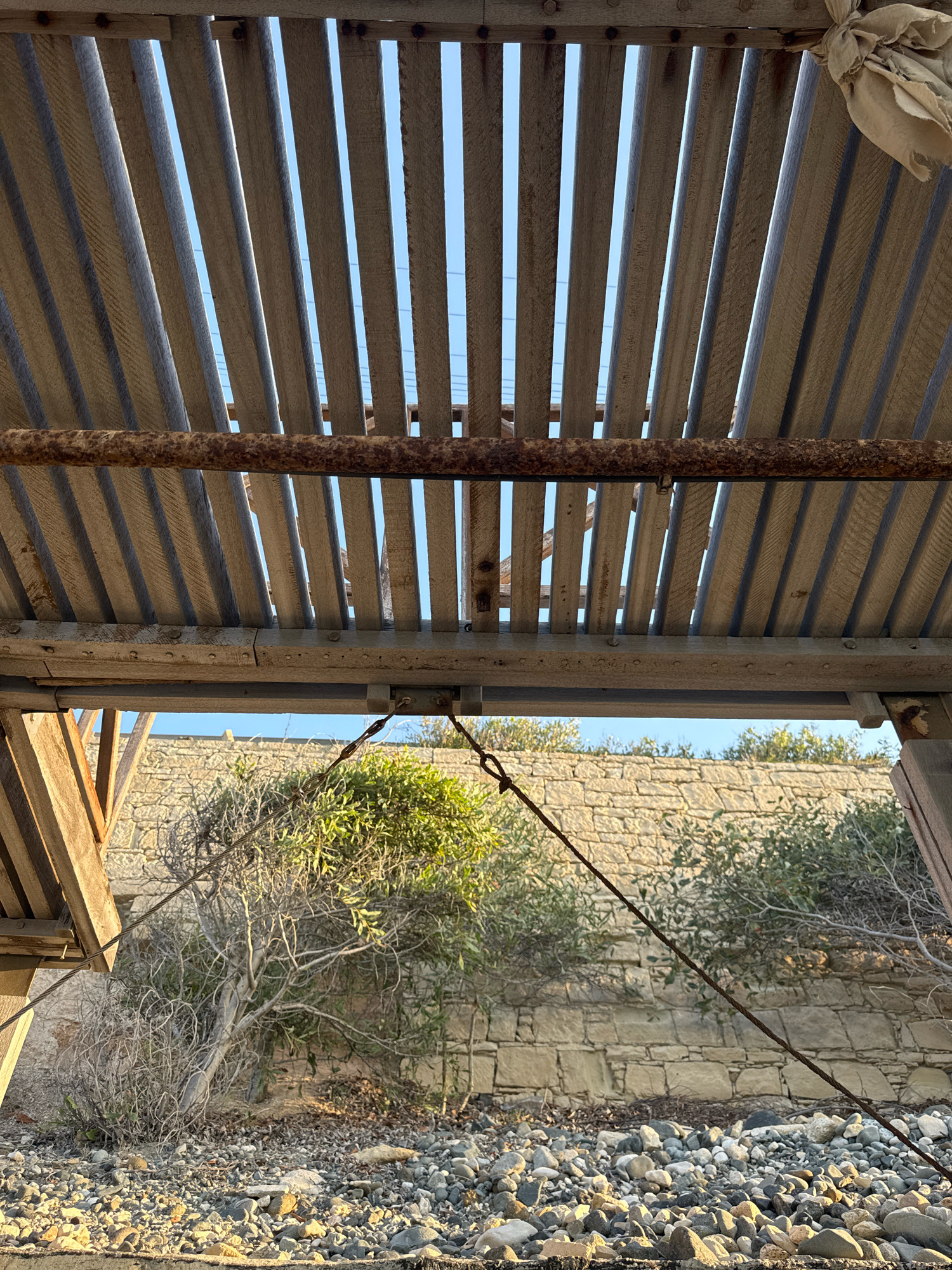 An outdoor scene in Agios Tychona Community, Cyprus, features a rustic wooden structure with a slatted roof, possibly a pergola or a simple shelter. The wooden slats, aged and weathered, allow sunlight to filter through, creating patterns of light and shadow. A rusty metal pipe runs horizontally beneath the roof. Below the structure, a stone wall forms the background, with vegetation and dry bushes growing in front of it. The ground is covered with pebbles and rocks. A thick wire cable stretches diagonally across the lower part of the frame, anchored to the structure on the right and disappearing out of frame to the left. The lighting suggests it is either early morning or late afternoon. No people are visible.