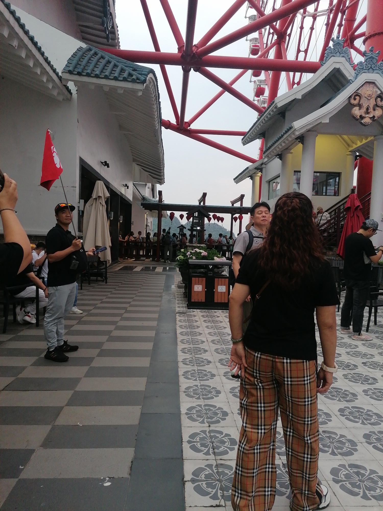 The image captures an outdoor scene in Hong Gai Ward, Vietnam, with a distinctive blend of architectural styles and human activity. Dominating the background is a large red Ferris wheel, its structure partially visible behind buildings designed with traditional Asian rooflines and ornamentation.
In the foreground, a woman stands with her back to the camera, wearing a black t-shirt and plaid trousers. To her left, a man holds a red flag with a white emblem. He is dressed in a black shirt, light-colored pants, and a baseball cap. The man is interacting with a few people sitting behind him. Further back, there's a group of people gathered on a raised platform, some standing and others sitting, observing the scene.
The ground is paved with a patterned tile design, transitioning from a gray and white checkered pattern near the flag-bearer to a floral motif closer to the woman in the foreground. The buildings on either side create a pathway leading towards the people in the distance. A few red lanterns can be seen hanging in the middle of the pathway.