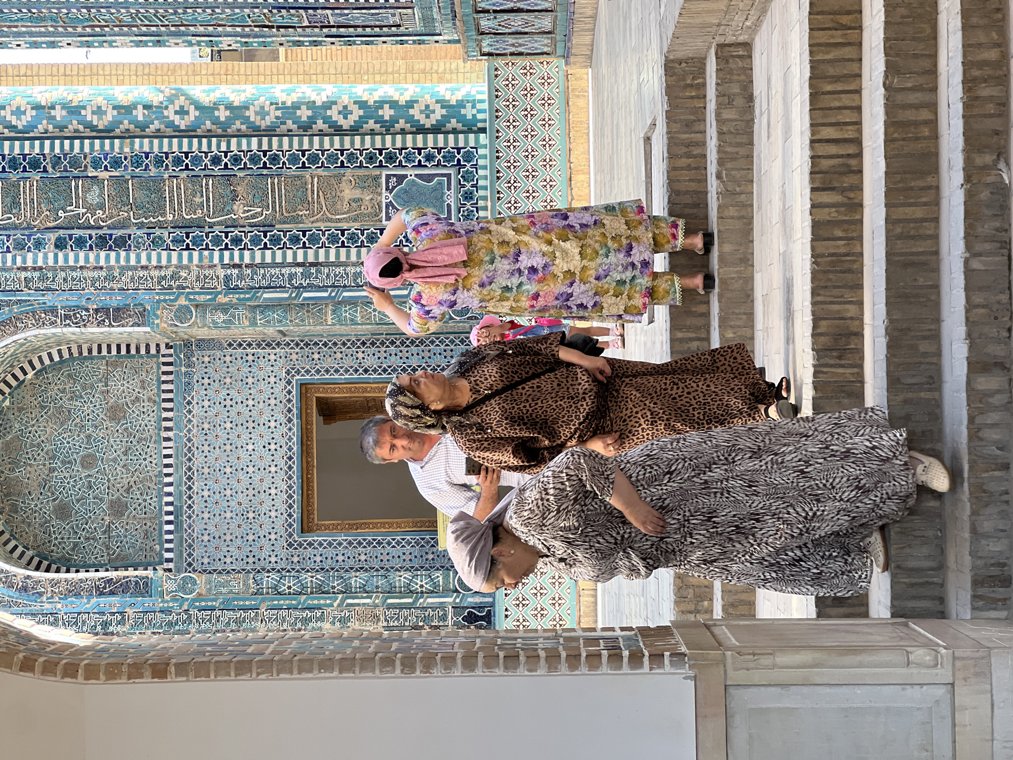 The image shows an outdoor courtyard with intricate blue and turquoise tilework on the walls of a building, likely a mausoleum, in Samarkand, Uzbekistan. In the foreground, four people are visible on a set of stone steps. On the left, a woman in a patterned grey and white dress and headscarf is looking down. Next to her, a woman in a leopard-print dress and headscarf is looking to the right. Behind them, a man in a white collared shirt is holding a smartphone and looking towards the right. Further back and to the right, a woman in a colorful floral dress and pink headscarf is posing with her hands on her head. A young girl in a pink dress is partially visible behind the woman in the floral dress. The overall atmosphere suggests a tourist visit to a historical and culturally significant site.