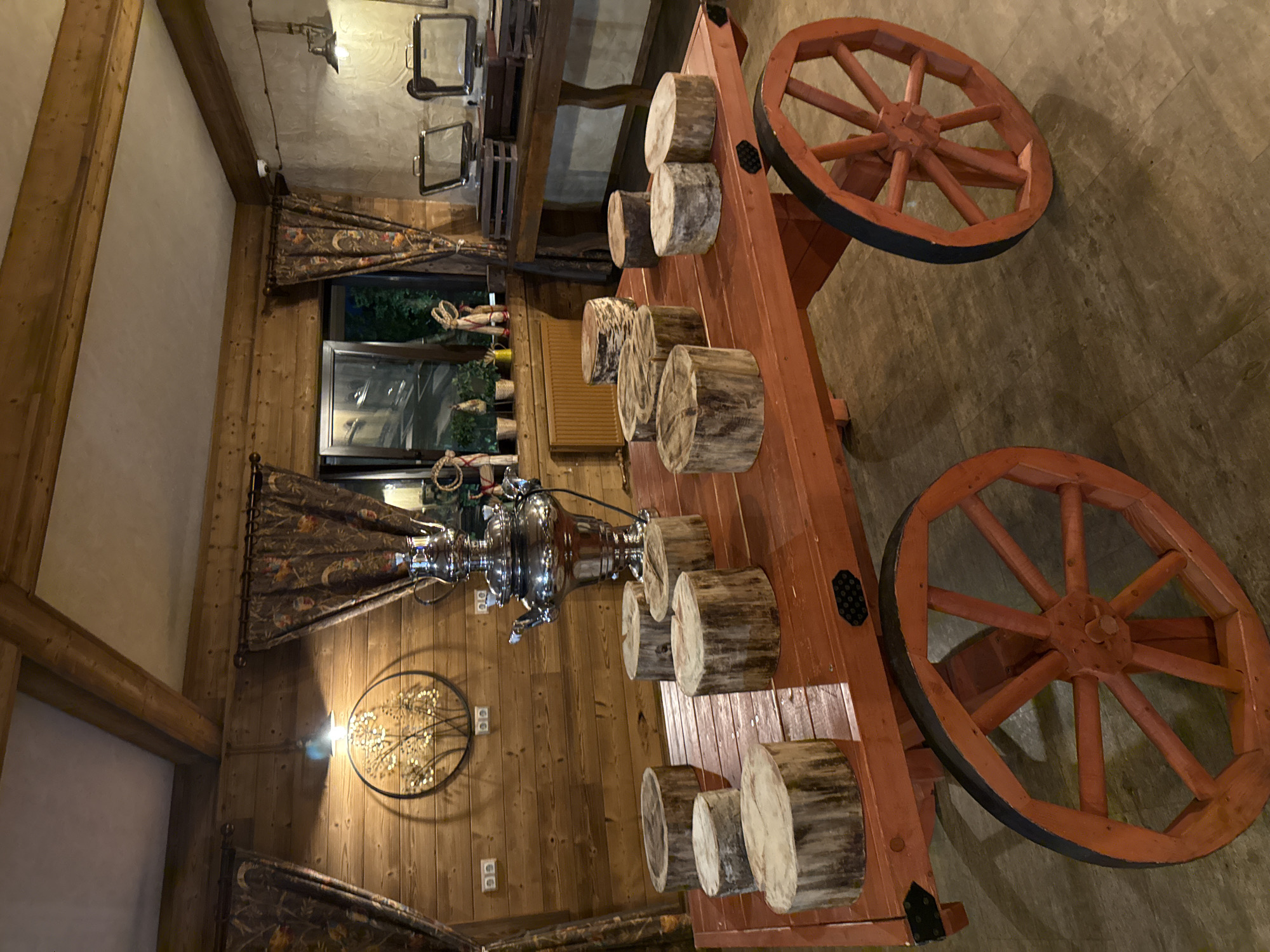 The image shows the interior of a restaurant or similar establishment in Peresvet, Russia. The focus is a rustic-style wooden table resembling a wagon. The table is decorated with cross-sectional cuts of logs and a samovar. The environment includes wooden walls, drapes, a window with a view of greenery, and decorative lighting fixtures. The overall impression is of a traditional Russian-themed dining space.