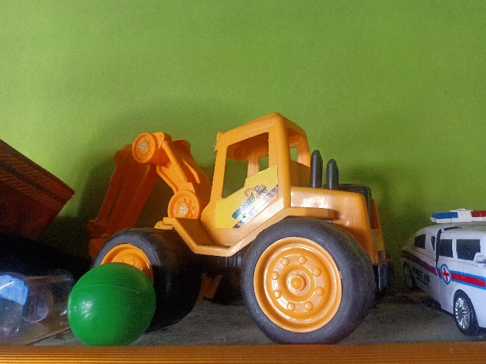 This media file, captured in Pubian, Indonesia, displays a still life arrangement of children's toys on what appears to be a shelf or flat surface, set against a plain, light green background.

The central and most prominent subject is a large, yellow plastic toy excavator. It features a detailed cabin with an open design, a long arm with a scoop (though the very end of the scoop is not fully visible), and four large wheels with orange rims and black tires. A colorful sticker or decal depicting what looks like construction vehicles is visible on the side of the excavator's cabin.

To the left of the excavator, partially obscured by its front wheel, is a smooth, bright green spherical toy ball. Another toy, possibly a dump truck or a similar vehicle, is partly visible on the far left, showing only a portion of its brown or orange bed.

To the right of the excavator, a white toy ambulance is visible. It has red and blue flashing lights on its roof, and a red cross symbol on its side, framed by blue and red stripes.

The objects are arranged horizontally across the frame, with the toys resting on a light brown or wooden surface that forms the bottom edge of the image. The green background appears to be a wall or a fabric surface, showing a slight texture. There is no discernible activity taking place, and no specific time of day or weather conditions can be inferred. No legible text is visible on any of the toys.
