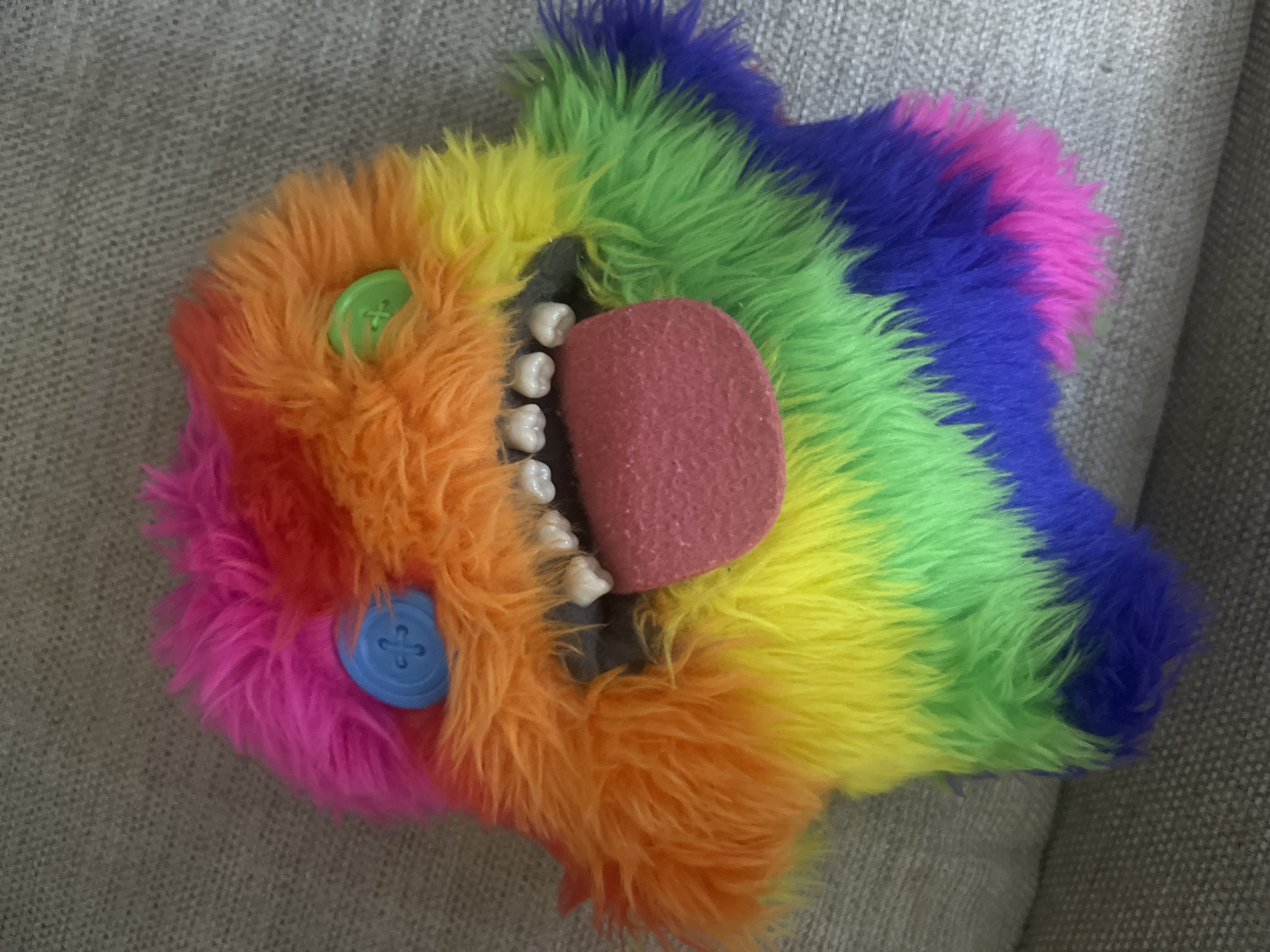 The image shows a close-up, top-down view of a colorful, plush monster toy with a rainbow striped body. The toy has two mismatched button eyes, one blue and one green, and a wide mouth with several white teeth and a pink tongue sticking out. The toy is resting on a grey, textured surface, which appears to be a couch or sofa. There is no indication of the city of Aktau, Kazakhstan, or any other specific location.