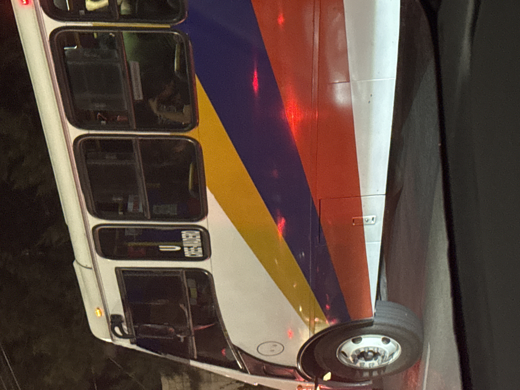 The image displays the side view of a public transportation bus at night, strongly suggesting a RUTA (Red Urbana de Transporte Articulado) bus from Puebla, Mexico, given its distinctive color scheme. The bus features a white body with prominent diagonal stripes in red, yellow, and blue extending along its length.
The scene is set after dark, as indicated by the deep shadows in the background and the artificial lights. Several bright red reflections are visible on the bus's colorful stripes, likely originating from an external light source or the bus's own lights. A small, illuminated amber marker light is also visible near the front wheel well. The front wheel, complete with a reflective silver hubcap, is clearly in view.
Through the bus's windows, which appear dark and reflective, faint shapes that could be passengers are discernible inside the vehicle. On one of the windows, the word "EMERGENCIA" (Spanish for "Emergency") is clearly visible in white text, printed on the interior and seen in reverse through the glass. This text, accompanied by an arrow, likely marks an emergency exit or instruction. The bus appears to be stationary or moving slowly in an urban environment.