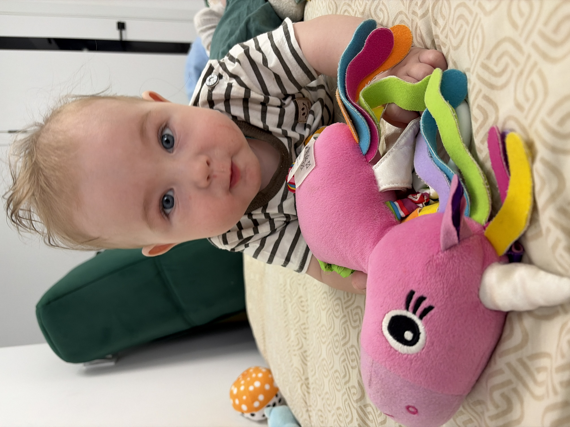 In a close-up, indoor shot, a fair-skinned baby with blonde hair, blue eyes, and pink cheeks lies on its stomach on a beige, patterned blanket. The baby is wearing a black and white striped t-shirt with a brown collar and a small circular emblem featuring a smiling face. The baby's hands are visible, resting on a pink plush toy shaped like a unicorn with a white horn and a black eye. The toy has a rainbow-colored mane made of fabric strips in shades of pink, orange, yellow, green, and blue. In the background, a dark green cushion or pillow is visible against a white wall, and part of a dark door is on the right. The scene is likely a home environment within the city of Chaikovsky, Russia.