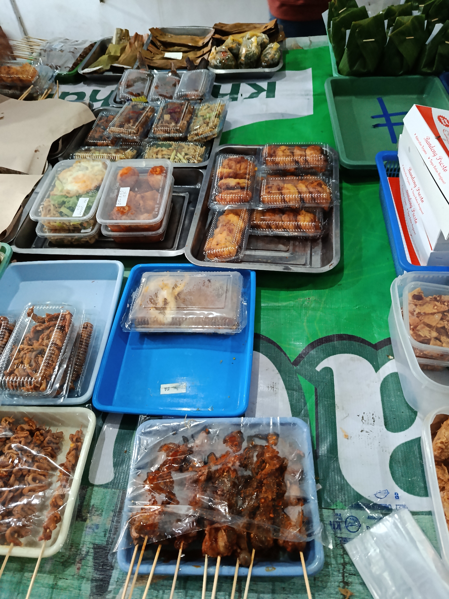 The image depicts a street food stall in Keboansikep, Indonesia. The stall features a variety of Indonesian dishes displayed in plastic containers and trays. Among the offerings are what appear to be fried snacks, grilled skewers of meat, and some wrapped food items. The food is arranged on a green tarp that serves as the stall's surface. In the background, more food items are visible, some wrapped in banana leaves, and a person's arm wearing a reddish-orange shirt is partially visible, suggesting they are tending to the stall. The lighting indicates it might be evening or an indoor setting. There is a white box with red and blue lettering, possibly indicating a brand or product name, in the upper right portion of the image.