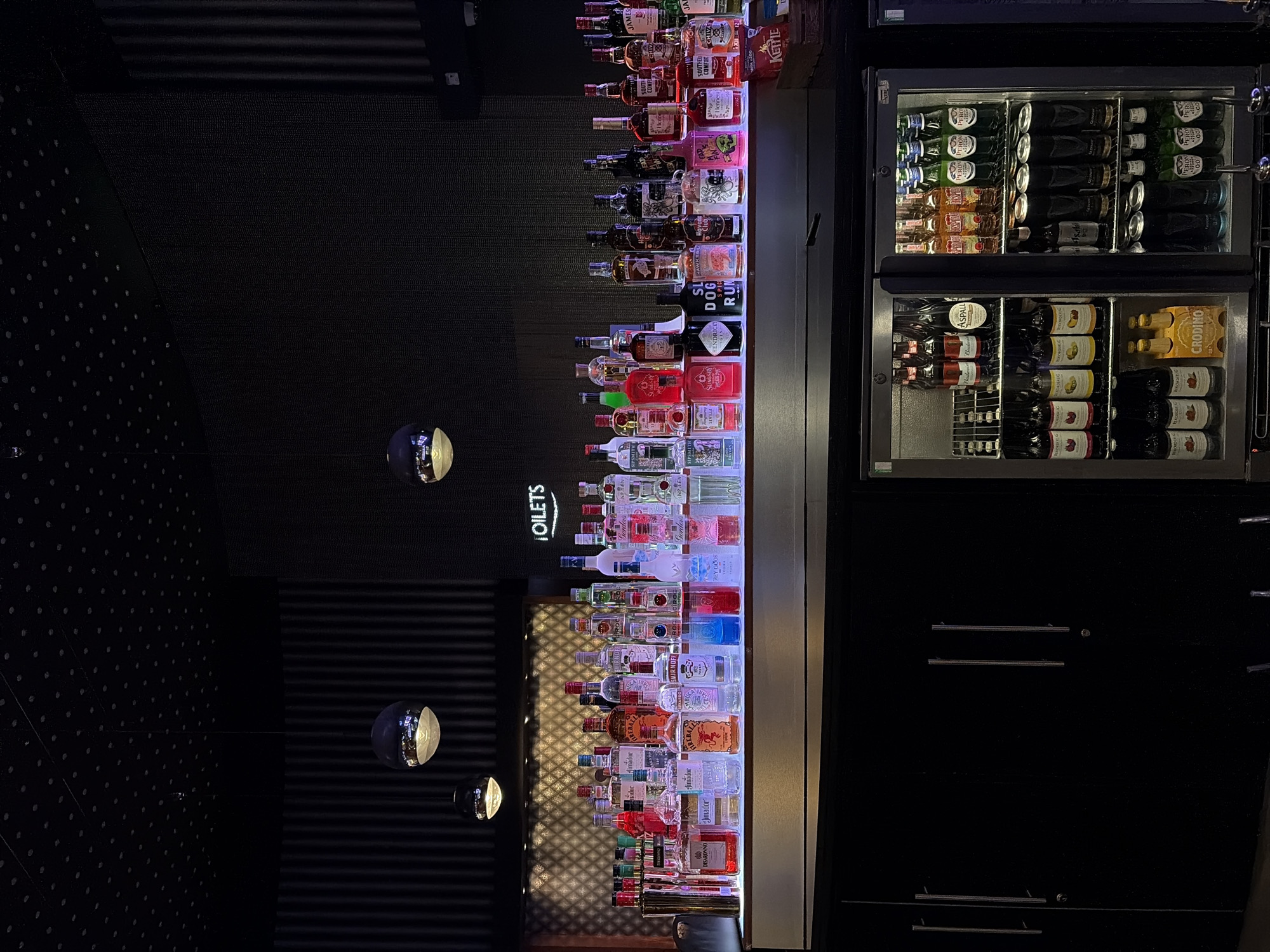 The image depicts a well-stocked bar display with numerous bottles of liquor and other beverages arranged on shelves. The bar appears to be located in the city of Manchester, United Kingdom, as indicated by the "TOILETS" sign in English. The bottles are illuminated from below by colored LED lights, creating a vibrant and attractive display. Behind the bar, a dark, textured wall is visible, and a few spherical pendant lights hang from the ceiling, casting a soft glow. A refrigerator unit is positioned below the bar, filled with various bottles and cans of drinks. The overall scene suggests a lively and modern bar or nightclub setting, likely busy with patrons, though no people are visible in the frame. The environment is clean and organized, with a focus on showcasing the beverage selection.