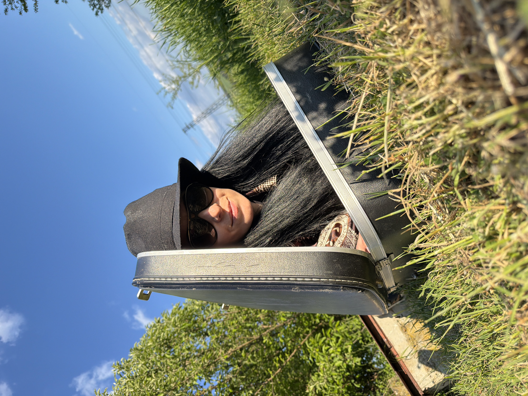 A woman wearing sunglasses and a hat is lying inside an open black suitcase. The suitcase is sitting on the ground with grass and trees visible in the background. The sky is blue with some scattered clouds. The exact location within Pleven, Bulgaria, cannot be determined from the image.
