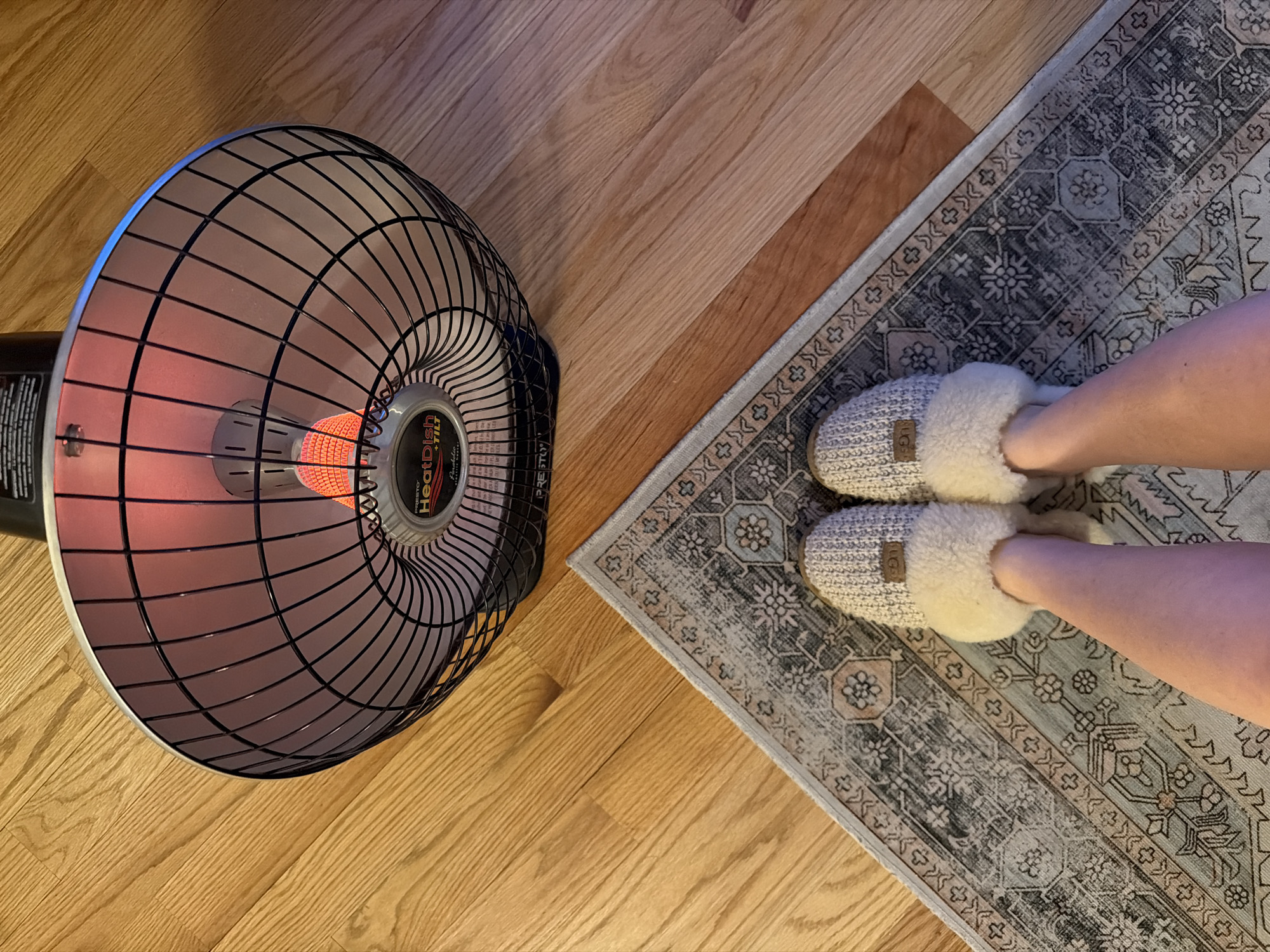 The image shows a top-down view of a section of a wooden floor, a small rug, a space heater, and a person's feet in slippers. The floor is made of light-colored wood planks. To the left, a round space heater with a black grill and a red heating element is positioned on the floor. To the right, a rectangular rug with a grey and white pattern is partially visible. At the top of the rug, a person's feet are wearing light-colored, fluffy slippers. The location is Morton Grove, United States of America.