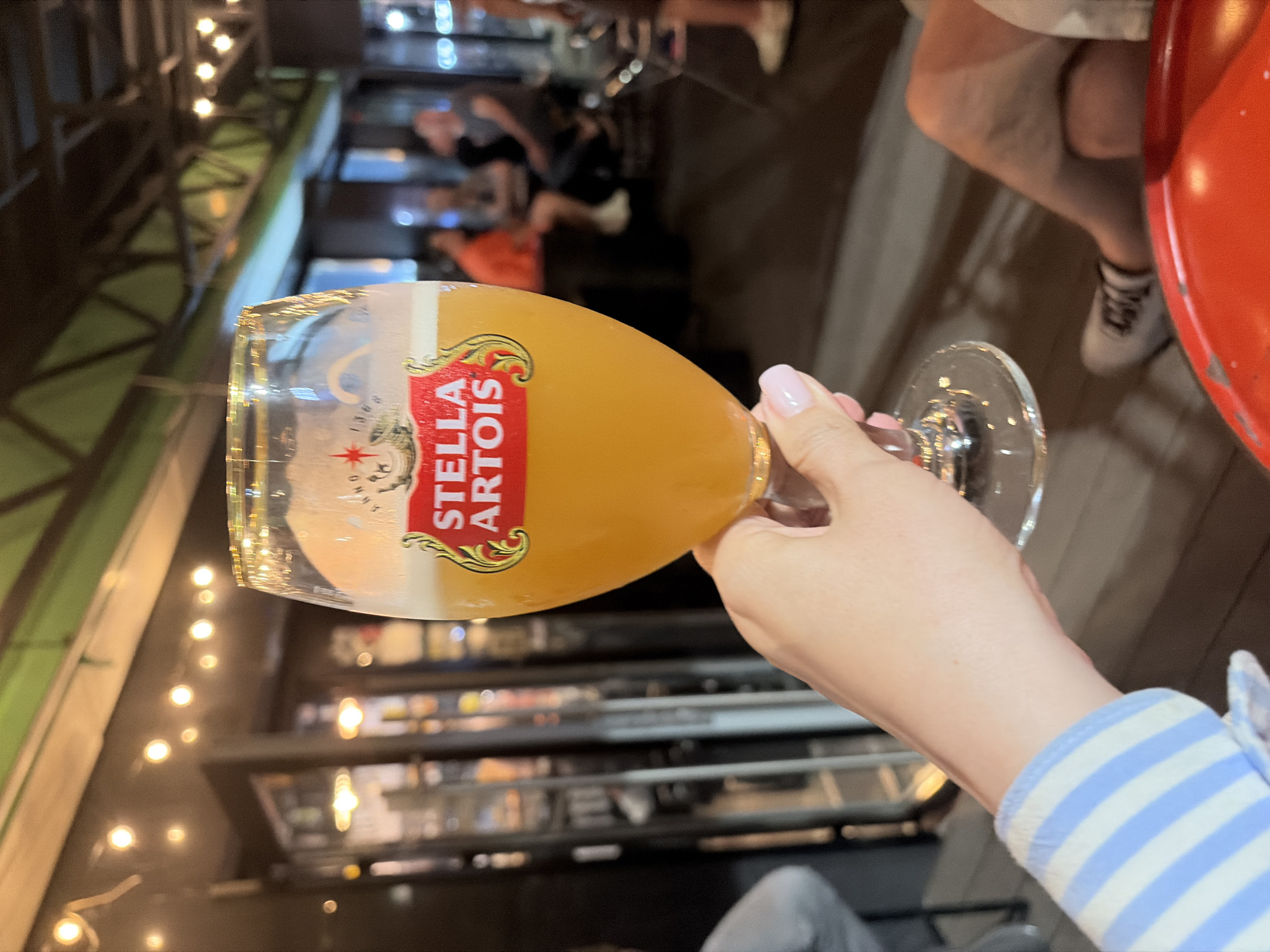 A person's hand, with neatly manicured pink nails, holds a glass of Stella Artois beer. The glass is positioned in the foreground, with the logo prominently displayed on its surface. The background reveals an indoor setting, possibly a bar or restaurant, with blurred figures of people seated at tables. String lights are visible overhead, casting a warm glow. The flooring appears to be made of wood. The environment suggests a casual social gathering. The location is not discernible from the image itself.