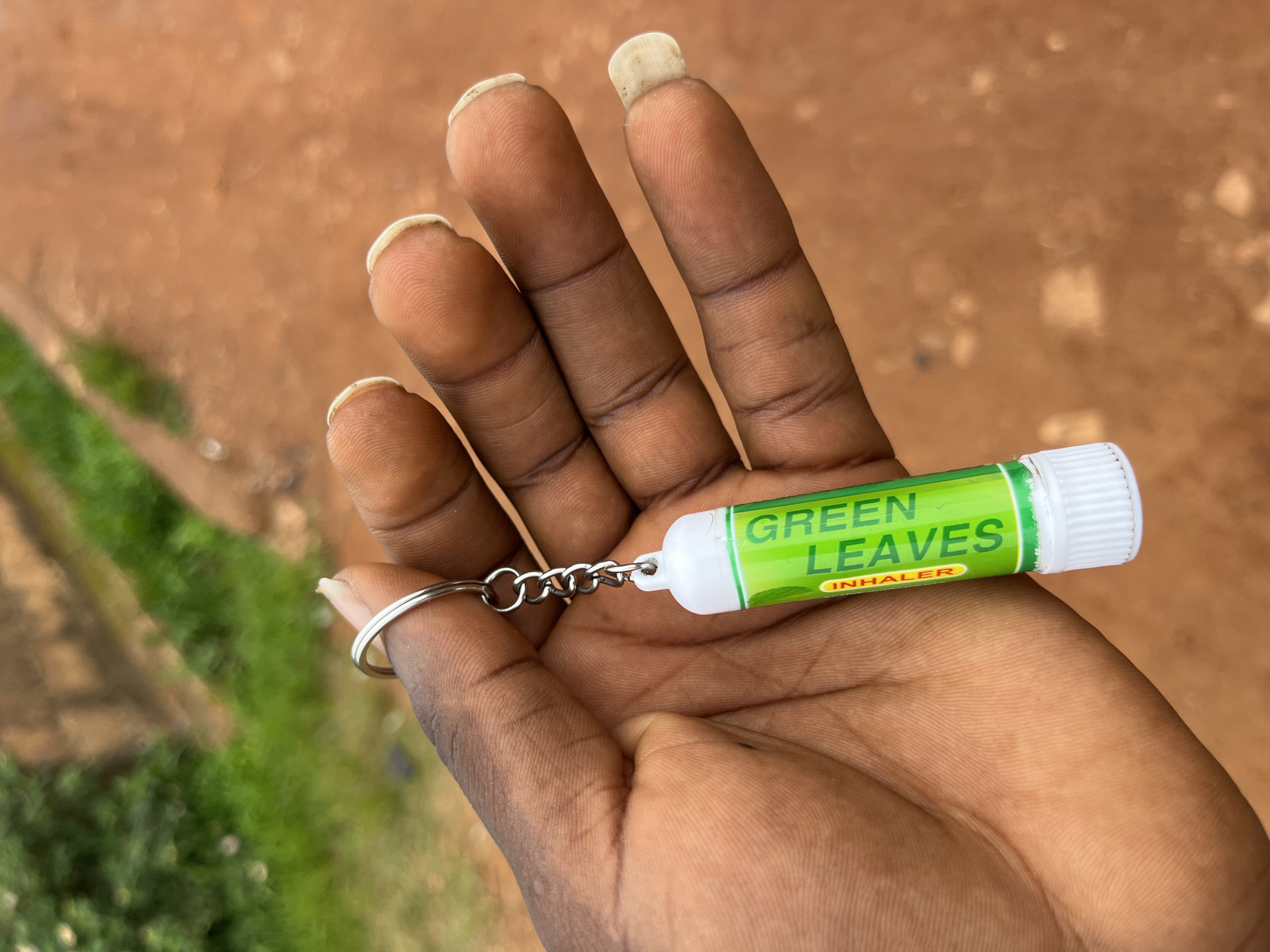 A person's dark-skinned hand is held up, displaying a green and white keychain inhaler. The inhaler has the words "GREEN LEAVES" and "INHALER" printed on it in white text. The hand is positioned in front of an out-of-focus background of reddish-brown dirt and green foliage, consistent with an outdoor environment in Bukuru, Nigeria. The person's fingernails are visible and appear unpolished and natural. The lighting suggests it is daytime.