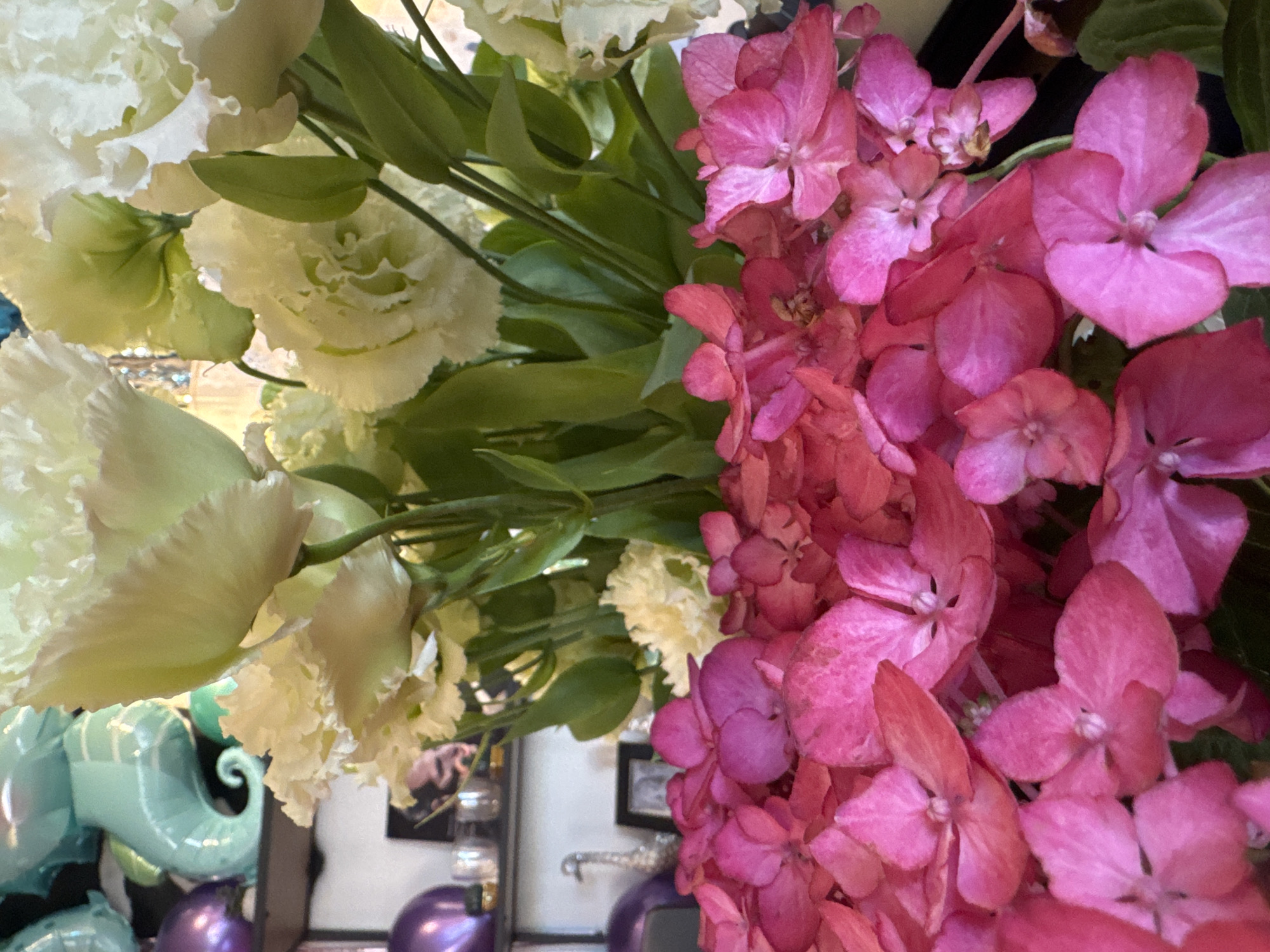 A close-up shot shows a bouquet of pink hydrangeas and white lisianthus flowers. The hydrangeas fill the foreground, with their delicate petals clustered together in varying shades of pink. Behind them, the white lisianthus blooms are partially visible, their ruffled petals creating a soft, ethereal contrast. The backdrop is blurred but appears to be an indoor setting with shelves holding decorative items, including sea-horse-shaped balloons and purple spheres. The image is well-lit, highlighting the textures and colors of the flowers. The scene is devoid of human presence and action, focusing solely on the floral arrangement and its immediate surroundings. No specific location in Kremenchuk, Ukraine can be determined from the visual information provided.