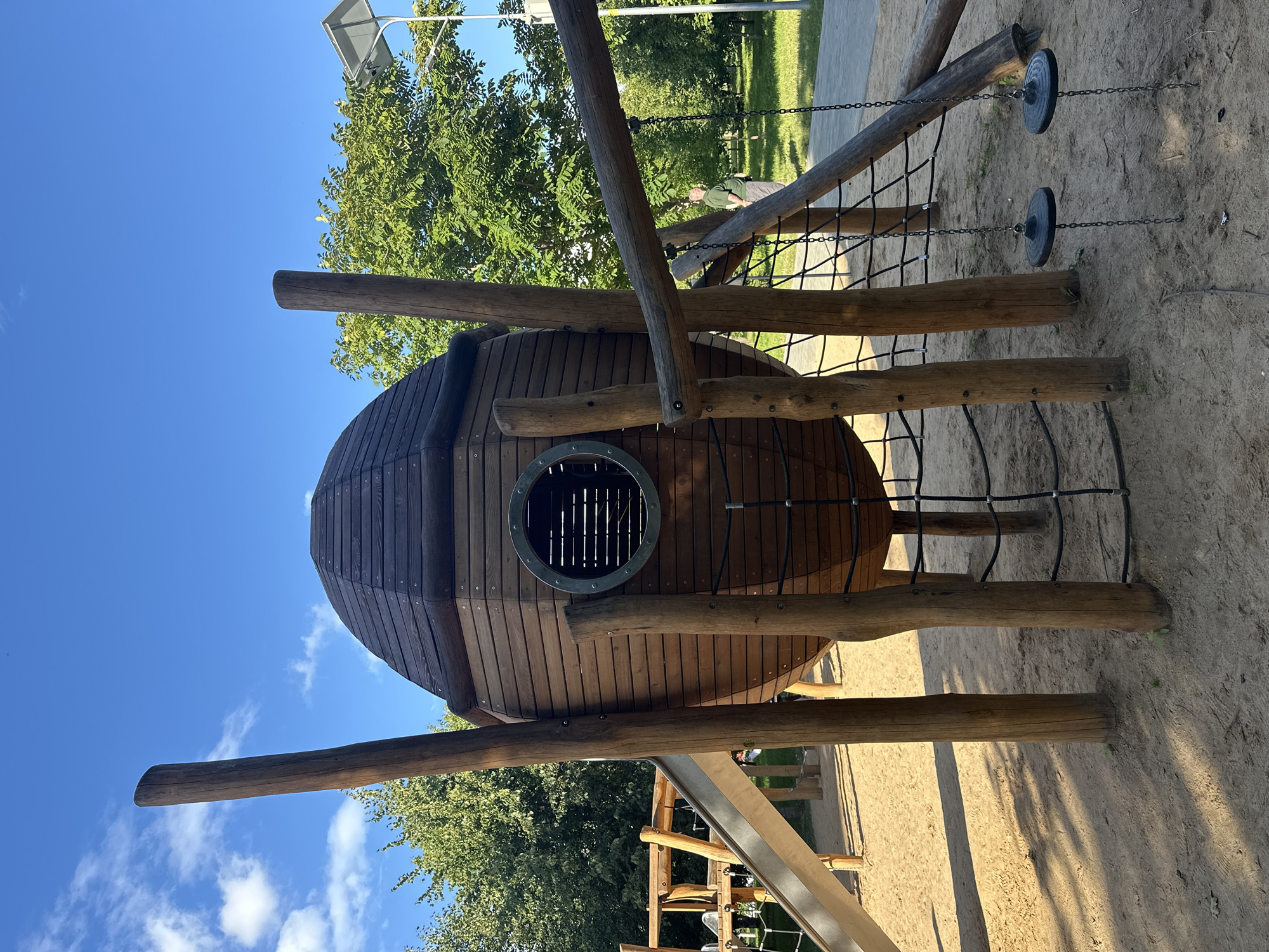 The image shows a playground with a large, egg-shaped wooden structure in the center. The structure has a circular window with a barred opening and is supported by thick wooden posts. Ropes and chains create a net-like climbing area around the base of the structure, and two circular swing seats hang from chains. To the left, a silver slide descends from a wooden platform. The ground is covered in sand. In the background, there are trees and a blue sky with some white clouds. A person is visible in the distance, standing on a paved path between the playground and a grassy area. The playground is located in Minsk, Belarus.