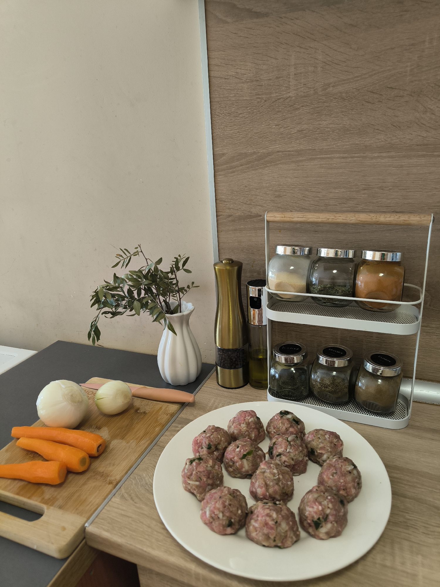 The image captures a close-up, elevated view of a kitchen counter showcasing ingredients and spices prepared for cooking. On the right side of the counter, a light wood-grain surface holds a white, two-tiered spice rack with a wooden handle. The rack contains six glass jars with silver lids; three are on the top shelf and three on the bottom. The spices include what appears to be garlic powder (top left), dried herbs (top middle), and a reddish powder, possibly paprika or chili (top right). On the bottom shelf, visible labels read "DILL," "SAVORY," and "CUMIN."
Next to the spice rack, a tall, gold-colored pepper grinder stands beside a clear glass bottle of olive oil. A white, fluted vase holding green leafy branches is positioned further left. Below the vase, the counter transitions to a dark grey surface. On this surface, a wooden cutting board displays two peeled white onions and three peeled orange carrots. A pink-handled knife rests on the cutting board.
In the foreground, on a white round plate, approximately fifteen raw meatballs are arranged. The meatballs appear to be made from ground meat mixed with visible specks of green herbs and possibly finely chopped onions. The background features a plain light beige wall on the left and a wooden panel or cabinet on the right. The scene suggests a daytime setting, focused on the preparation of a meal. No people are visible in the image.