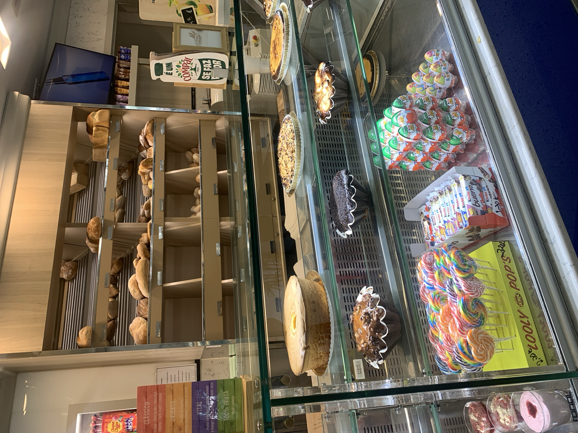 This image showcases a display case in what appears to be a bakery or confectionery shop in Odivelas, Portugal. The display is filled with a variety of baked goods, including loaves of bread, tarts, and individual cakes. Prominently featured are colorful lollipops arranged in a box labeled "LOOLY POP'S," as well as several packages of Kinder chocolate products. Various brands and product names are visible in the text, such as "Chupa Chups," "BEST&," "CLÁSSICO," "ORIGENS," "VITAL," and "COMPAL." The overall scene suggests a commercial establishment stocked with sweets and baked items. The lighting indicates it is daytime, and the orderly arrangement of products points to a well-maintained shop.