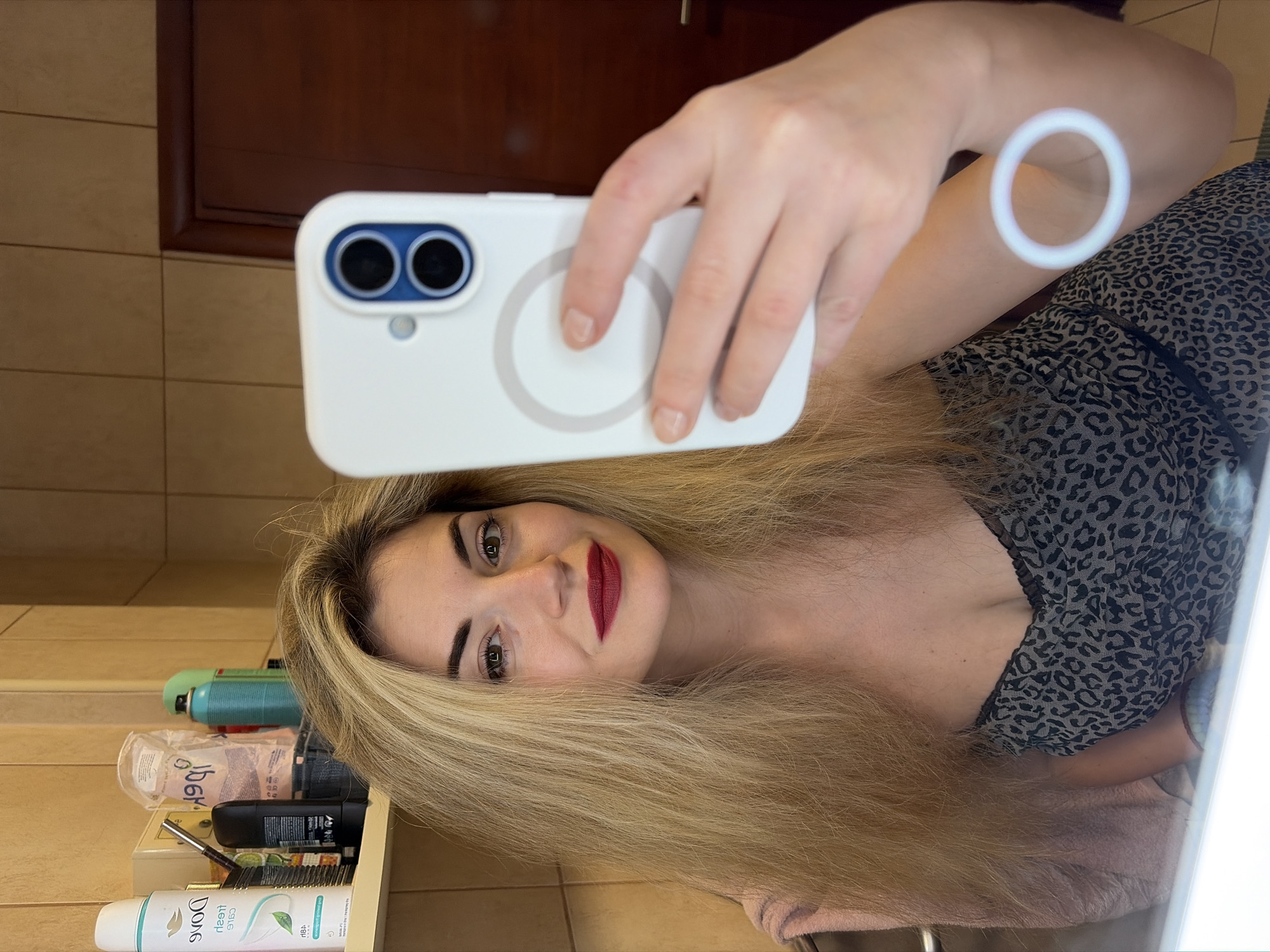 The image depicts an adult female subject capturing a mirror selfie within a bathroom, located in Budva, Montenegro. The female has long, blonde hair, brown eyes, and is wearing red lipstick. She is dressed in a black and gray leopard-print top with a V-neckline. Her right hand (viewer's left) holds a white smartphone, fitted with a white case featuring a prominent circular magnetic ring on its rear, pointed towards her reflection. A pink textile is partially visible over her left shoulder (viewer's right). The background consists of light brown square wall tiles and a white wall-mounted shelf. On this shelf, multiple personal care products are present, including an inverted 'Dove Fresh Care' spray deodorant, a black deodorant stick, and additional containers. A dark wooden door with a metallic handle is reflected behind the smartphone. A distinct white circular light reflection is visible on the subject's right bicep.