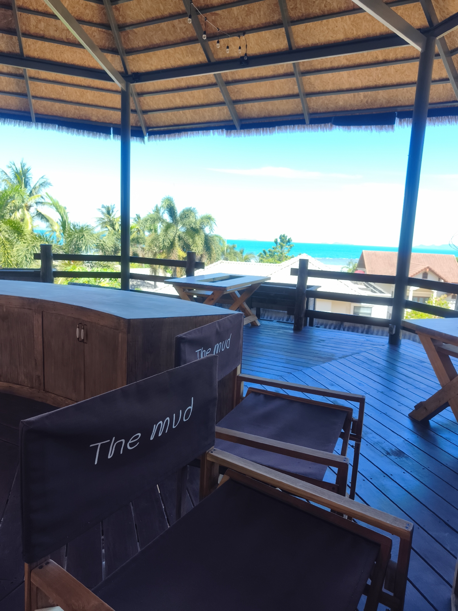 An outdoor covered seating area with a thatched roof overlooks the ocean in Baan Thong Po, Thailand. The foreground shows two dark canvas director's chairs with "The mud" written in white script on their backs. Behind the chairs, a curved wooden counter or bar is visible with two white drawer pulls. The floor of the seating area is made of dark wood planks. In the middle ground, there are several wooden tables with X-shaped legs and a wooden railing. Beyond the railing, palm trees and tropical vegetation are abundant, with houses visible in the distance. The bright blue ocean and sky form the background. The scene appears to be a scenic viewpoint or a restaurant/bar area. There are no people visible in the image.