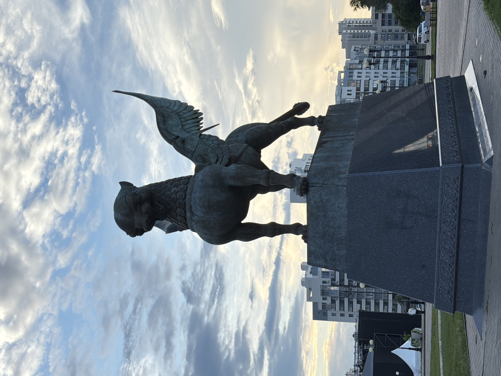The image shows a bronze statue of a winged lion, possibly a griffin, mounted on a large, dark stone pedestal. The statue is positioned on the right side of the frame, facing left, with its wings spread. Its body is powerfully built, with muscular legs and a detailed mane. The sky behind the statue is a mix of blue and white clouds, with a pale yellow glow on the horizon, suggesting it is either early morning or late afternoon.
In the background, there are modern residential buildings and streetlights. The architecture appears to be typical of a city in Russia, specifically Kazan, as indicated by the location context. A paved walkway and a grassy area are visible in the foreground and mid-ground. There are no people clearly depicted in the image, though there might be distant figures or cars, but they are not distinct enough to describe. The overall mood is somewhat serene, with the majestic statue set against a diffused sky. The time of day implies a calm atmosphere, perhaps before or after the day's peak activity. There is no visible text in the image.