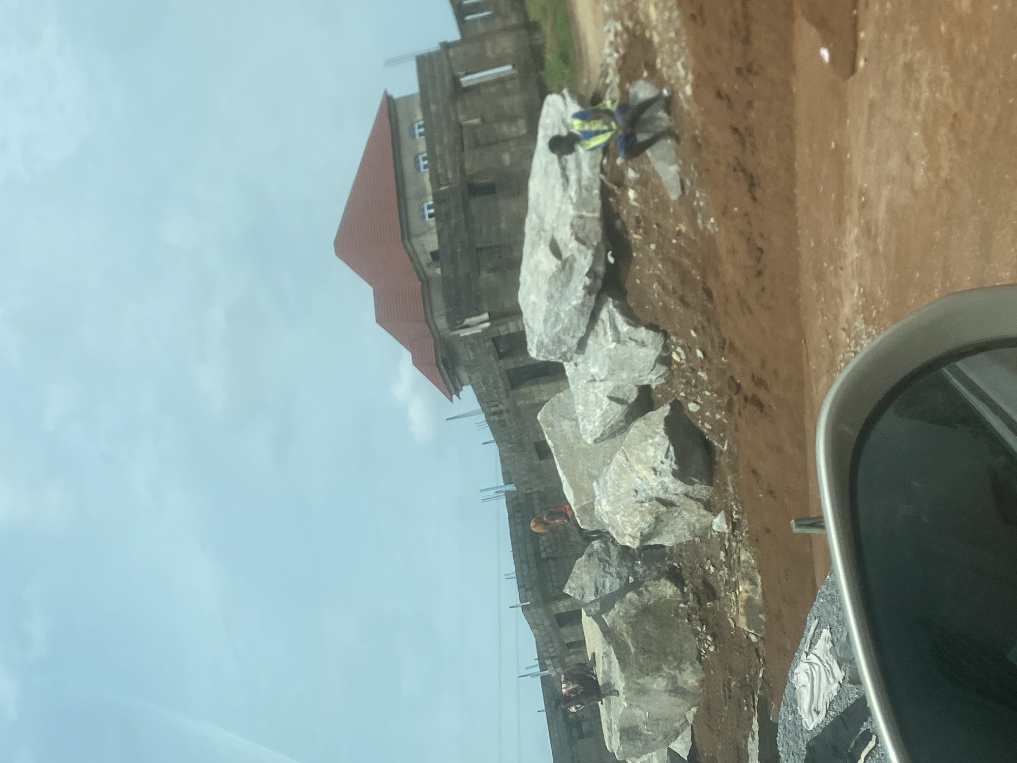 This image captures an active construction site in Gosa, Nigeria, under a bright, overcast sky. The perspective appears to be from inside a vehicle, with a portion of a car's side mirror visible in the lower right corner.
The central focus is a multi-story building in the early stages of construction, positioned on the left side of the frame. It features a prominent red-tiled roof at the top, while the main structure below consists of concrete and visible rebar. Several open window frames are visible on the building's facade.
In the foreground and mid-ground, a row of large, gray rocks of varying sizes runs across the scene, indicating construction materials. Several workers are present on the site. One worker, wearing a blue shirt and a high-visibility yellow-green vest, stands near these rocks. Further back, closer to the building, another worker is visible in a red or orange top, possibly wearing a hard hat. A third, less distinct figure can be seen near the base of the building on the left.
The ground is a mix of reddish-brown dirt and soil, characteristic of construction environments, with smaller debris scattered around. The bright, even lighting suggests it is daytime. No explicit text is clearly visible in the image.
