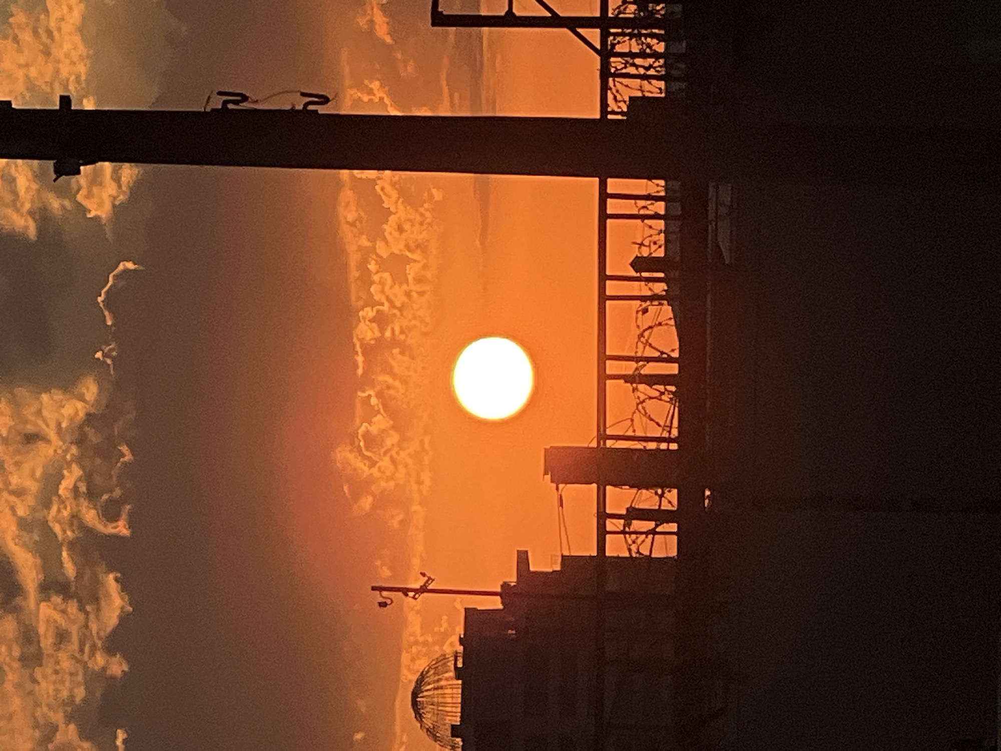 This is a photograph of a sunset viewed through an urban landscape. The sun, a bright, white orb, is positioned in the center of the frame, casting a warm, orange glow across the sky and illuminating scattered clouds. The scene is framed by dark, silhouetted structures, suggesting a cityscape. In the foreground, a horizontal beam, possibly a power line support, runs across the top of the image with two curved metal pieces attached. Below this, to the left, are wispy cloud formations. The middle ground is dominated by the silhouette of buildings, one with a domed structure at its base. A distinct element of this building is a horizontal bar with a red object attached, resembling a directional signal or antenna. To the right of the sun, another building's silhouette is visible, featuring a fence with barbed wire across its upper section, possibly indicating a secured area. The overall impression is one of an industrial or urban environment at dusk. The time of day is clearly sunset due to the low sun and warm, orange hues. The location is suggested to be Krasnodar, Russia. No text is visible in the image.