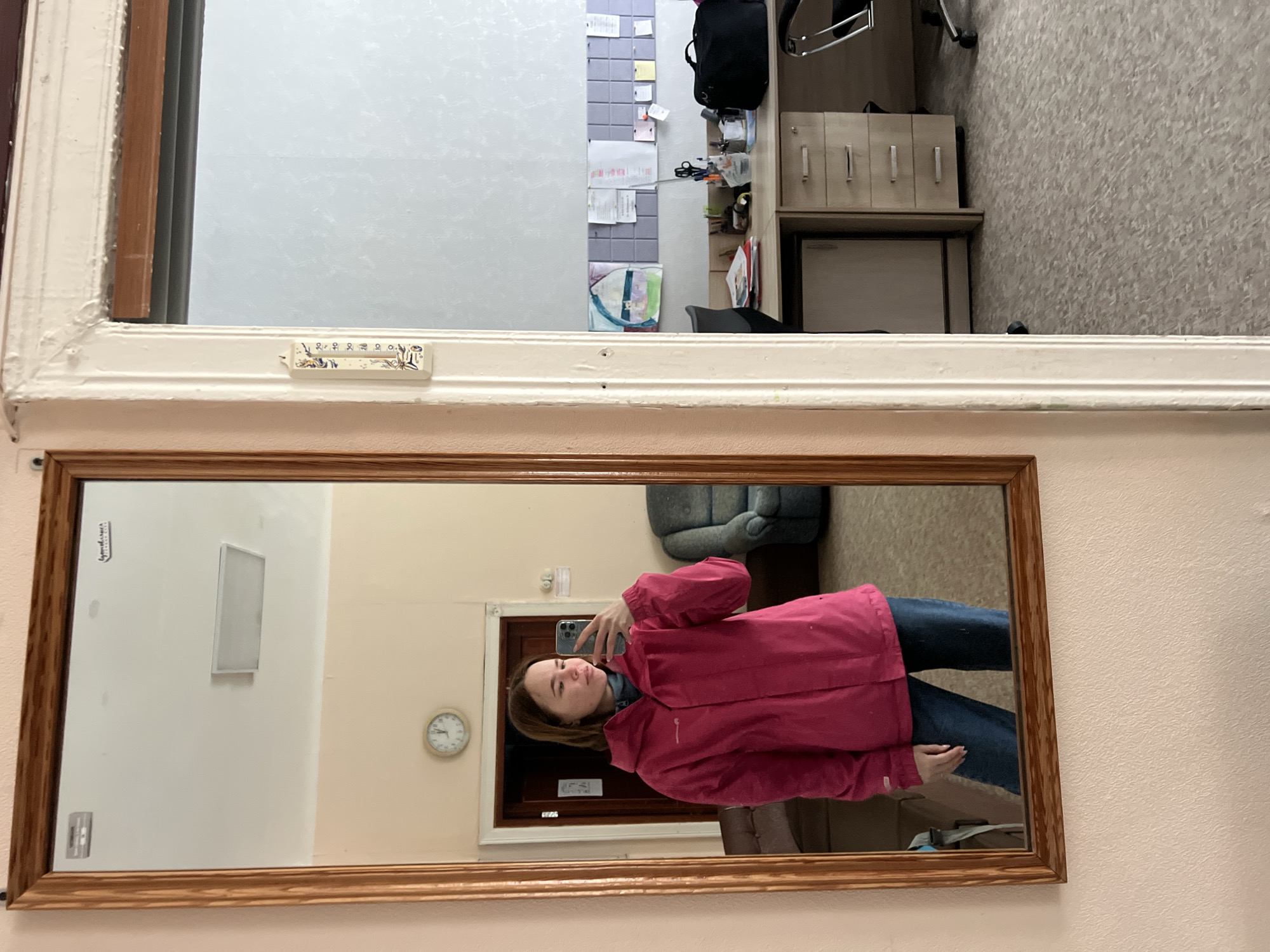 A woman in a pink jacket and jeans is taking a selfie in a mirror. The mirror has a wooden frame. Above the mirror is a window. The reflection in the window shows an office space with a desk, chair, filing cabinet, and bulletin board. A thermometer is attached to the window frame. In the mirror's reflection, a wall clock and a closed wooden door are visible. Based on the location information, the photo was taken in Chaikovsky, Russia.