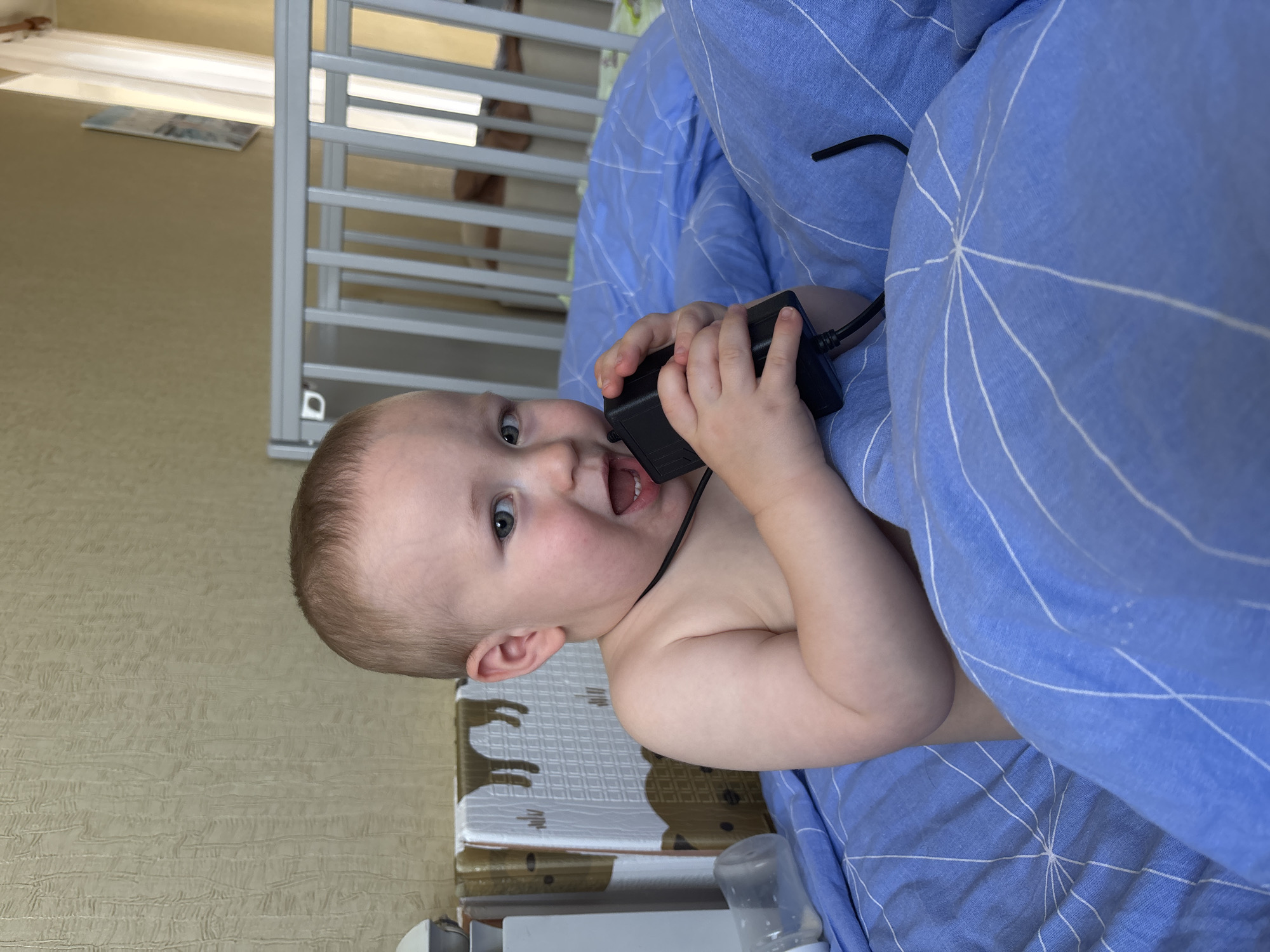 An infant with fair skin and light brown hair is lying on a bed with blue bedding. The infant is looking at the camera with an open mouth and is holding a black electronic device to its face. The infant is wearing a black cord around its neck. The background shows a crib and a wall with textured wallpaper. The scene appears to be indoors, likely in a residential setting in Naberezhnye Chelny, Russia.