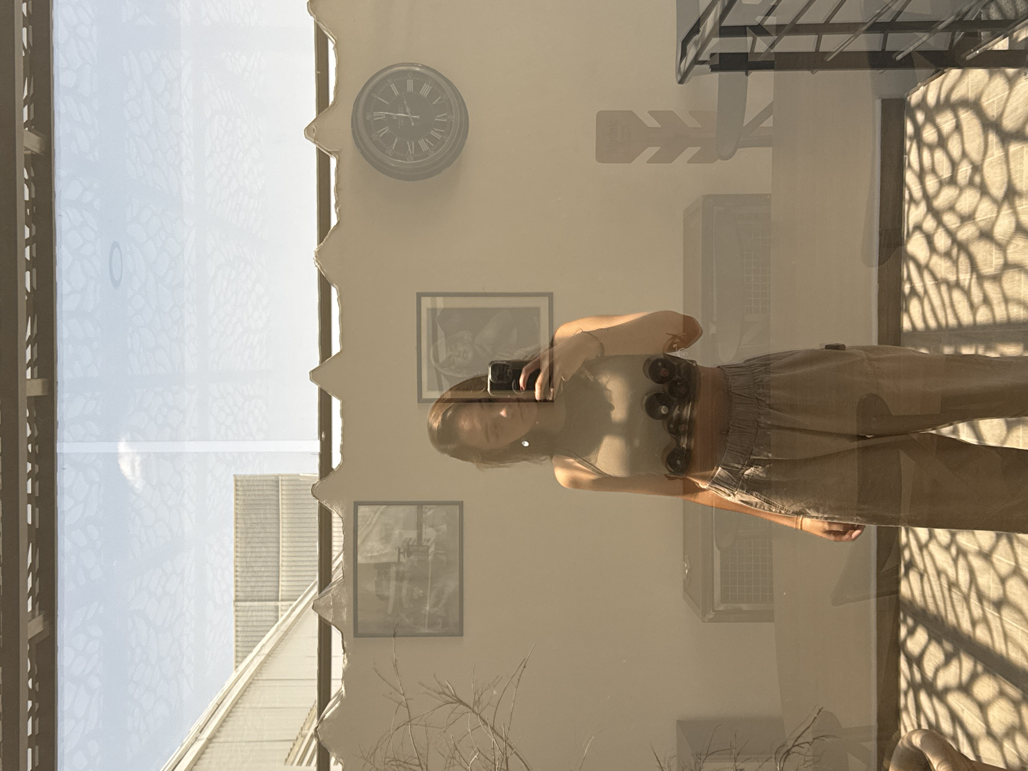 A woman with long brown hair is taking a selfie in a room in San Bernardino Tlaxcalancingo, Mexico. The room has a wall clock, two framed pictures, and an air conditioning unit. Light comes through a window with a patterned design.