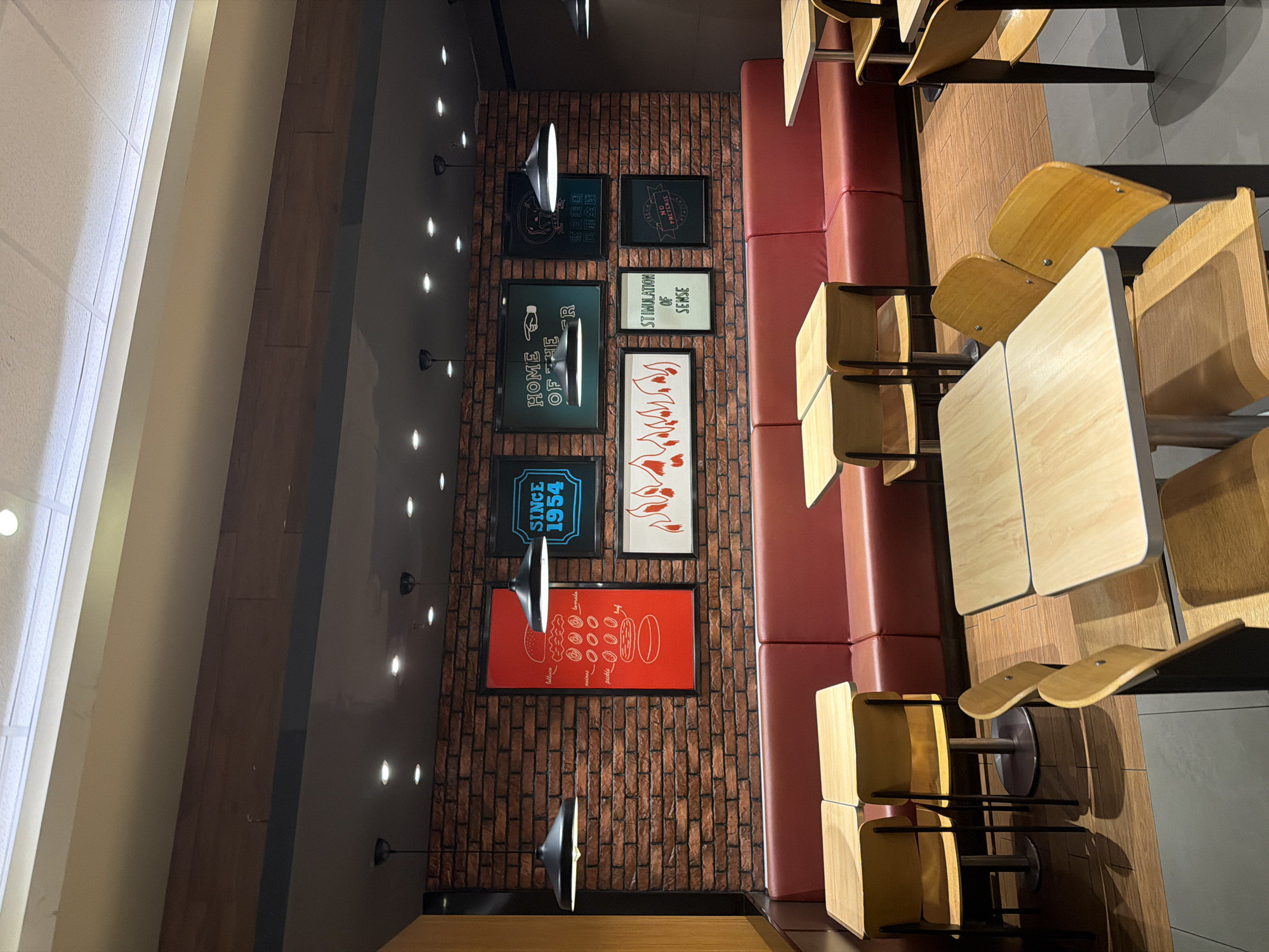 The image depicts the interior of a restaurant, likely located in the Jiangning District of China, evidenced by the inclusion of a phrase in Chinese that translates to "Originated in the United States, Popular around the World". The restaurant features a brick wall adorned with framed posters including one with the text "SINCE 1954" and "HOME OF THE ", another with a list of food components, and others with slogans. A built-in red bench seating runs along the wall adjacent to a row of wooden tables and matching chairs. Above the wall, a series of small spotlights are installed. The scene appears to be a quiet moment in the establishment, absent of people.
