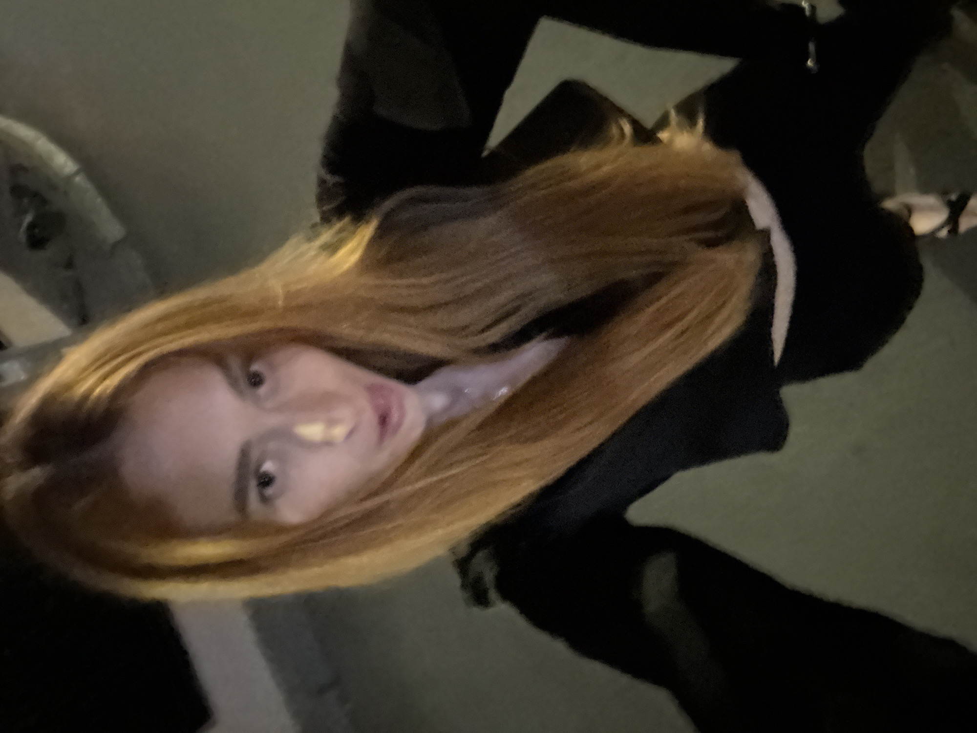 This image captures a close-up, rotated view of a young woman, likely a selfie, in Budva, Montenegro. The primary subject is a woman with long, straight, light brown to blonde hair that frames her face. She is looking directly at the camera with a neutral expression.
She appears to be wearing dark clothing, possibly a jacket or top, with a contrasting lighter, possibly white or off-white, trim or inner layer visible around her neck and chest area. A small metallic detail, possibly a zipper pull or clasp, is subtly visible on her dark clothing.
The setting appears to be dimly lit, suggesting it is nighttime or an indoor environment with low ambient light. Her face and hair are illuminated by a direct light source, possibly a camera flash or a nearby artificial light, creating some highlights and reflections. The background is dark and out of focus, offering minimal detail beyond blurry shapes that suggest an indistinct structure or environment. There are no clear identifying objects or specific location cues within the blurred background to confirm the exact location within Budva.
There is no visible text in the image.
