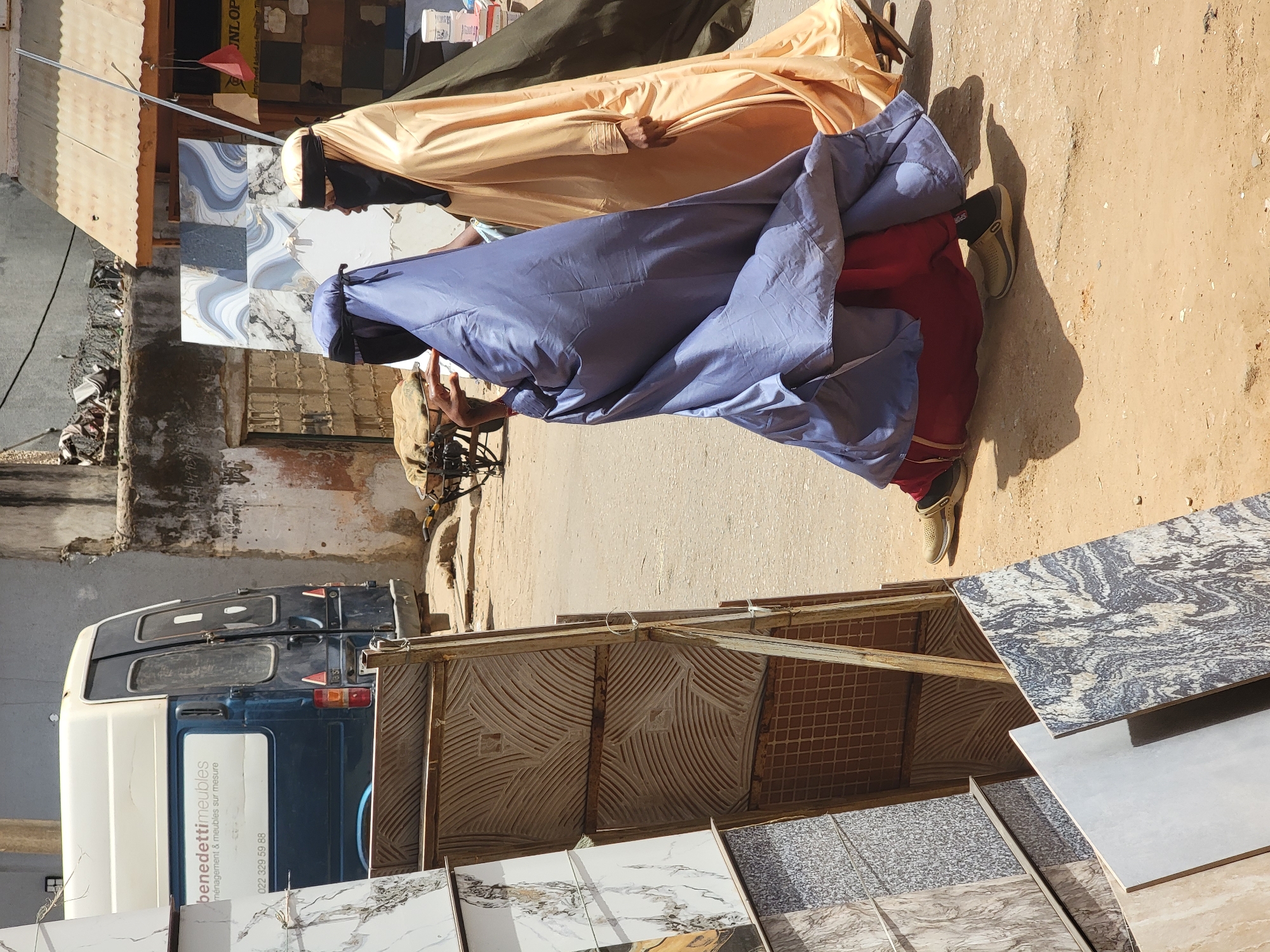 The image shows a street scene in Maiburiji, Nigeria. Two men dressed in traditional robes are walking on the street. One is wearing a pale yellow robe, and the other is wearing a blue robe over red pants. A van with the words "benedetti meubles" is parked nearby. There are also ceramic tiles displayed on a rack.