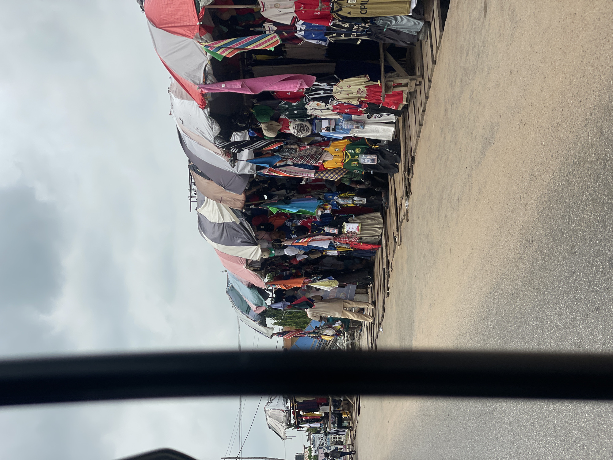 This image depicts an outdoor market scene along a paved road in Lokoja, Nigeria. A row of stalls, covered by colorful umbrellas and tarps, lines the right side of the road, displaying a variety of clothing items including t-shirts, jerseys, shorts, and other apparel. The merchandise is hung and displayed on wooden structures. Several people are visible within the market area, with one individual in the foreground wearing a light-colored traditional outfit. Another person can be seen in the distance on the left side of the road. The sky is overcast, suggesting a cloudy day. The foreground is partially obscured by a dark, vertical element, possibly part of a vehicle or structure the photograph was taken from. The overall atmosphere suggests a bustling local marketplace.