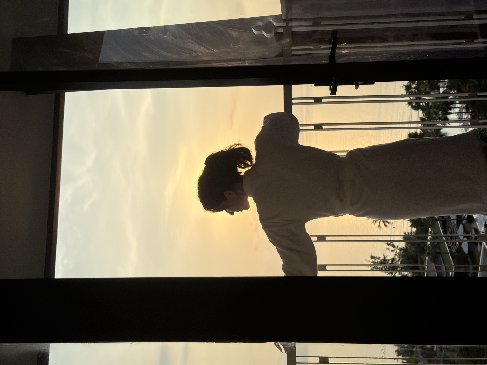 An individual, appearing female, is silhouetted against a golden sky, standing on an outdoor balcony in Dong Ninh Hoa Ward, Vietnam. The person is viewed from behind and slightly to the right, wearing a light-colored, knee-length terry cloth bathrobe. Their hair is tied back in a ponytail. They are facing left, looking out towards the horizon over a calm body of water, which reflects the warm, yellow-orange hues of the sunrise or sunset. Both forearms are resting on a light-colored metal railing, which features vertical and horizontal bars. The scene is framed by dark vertical elements of an open doorway or window from an interior room. The right frame includes a textured, possibly marble, panel and a dark door handle mechanism. Below the balcony, lush green foliage, specifically palm trees, is visible, along with multiple swimming pools and white sun umbrellas, indicating a resort or hotel complex.