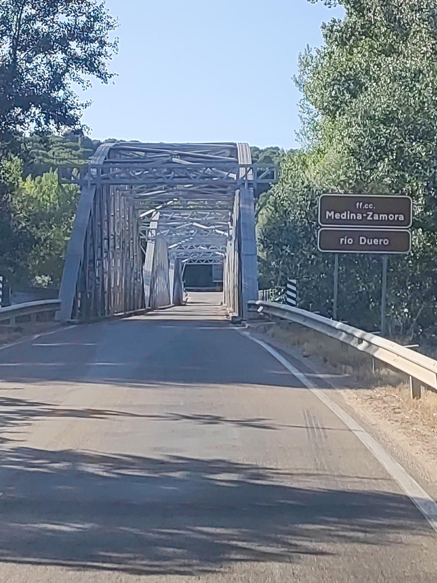 The image shows a view from a road approaching a steel truss bridge. The bridge spans over a river, identified by a sign as "río Duero." Another sign indicates "ff.cc. Medina-Zamora," likely referring to a railway line. The road is asphalt with white lane markings and a metal guardrail on the right side. The environment is rural, with trees and vegetation on either side of the road and surrounding the bridge. The sky is clear and blue. The lighting suggests it is daytime.