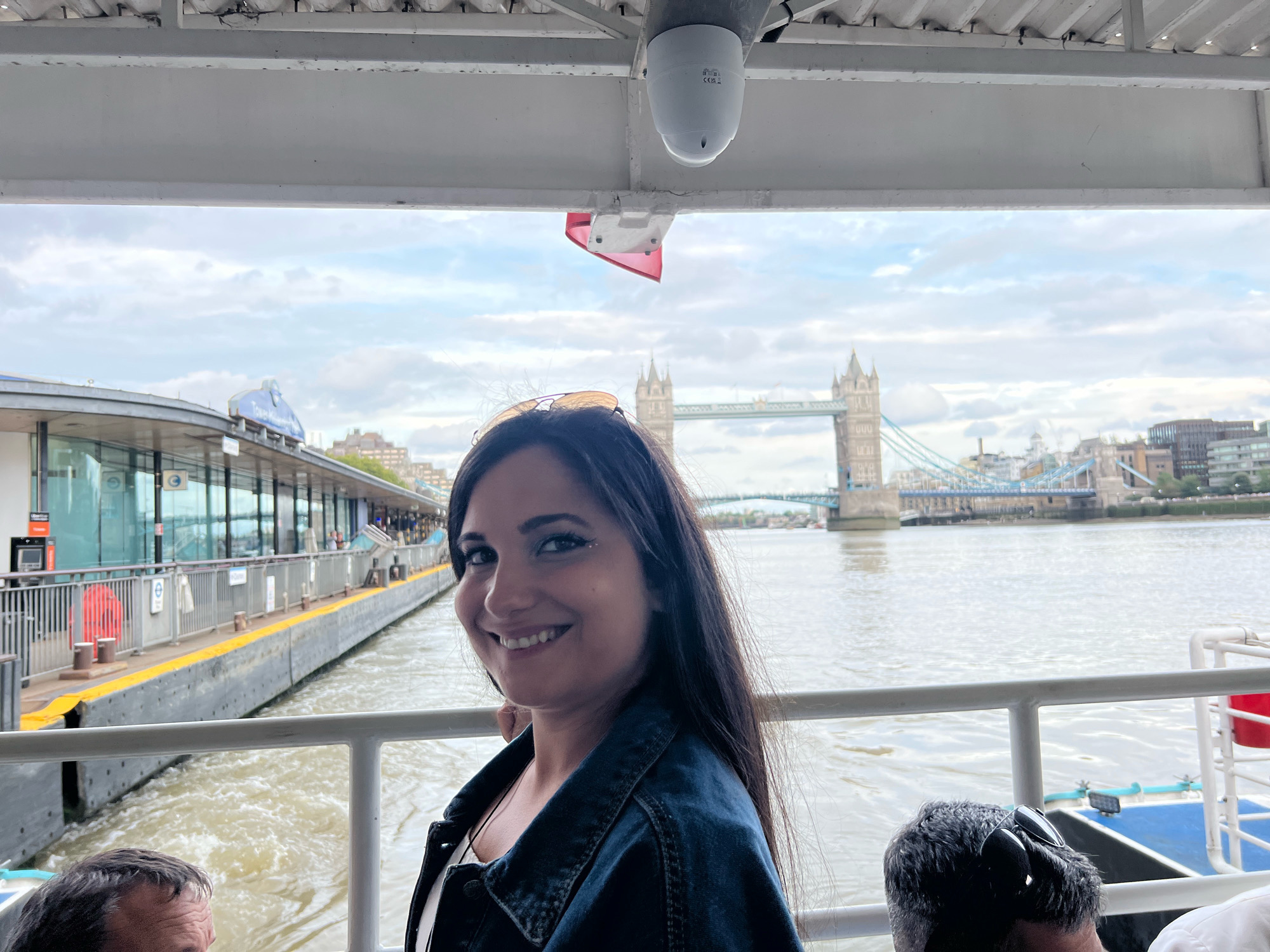 The image shows a woman with long dark hair and sunglasses perched on her head, smiling at the camera. She is on a boat with a white railing. In the background, Tower Bridge is visible over the River Thames, along with buildings lining the riverbank and a dock with a yellow line on the edge. The sky is overcast.