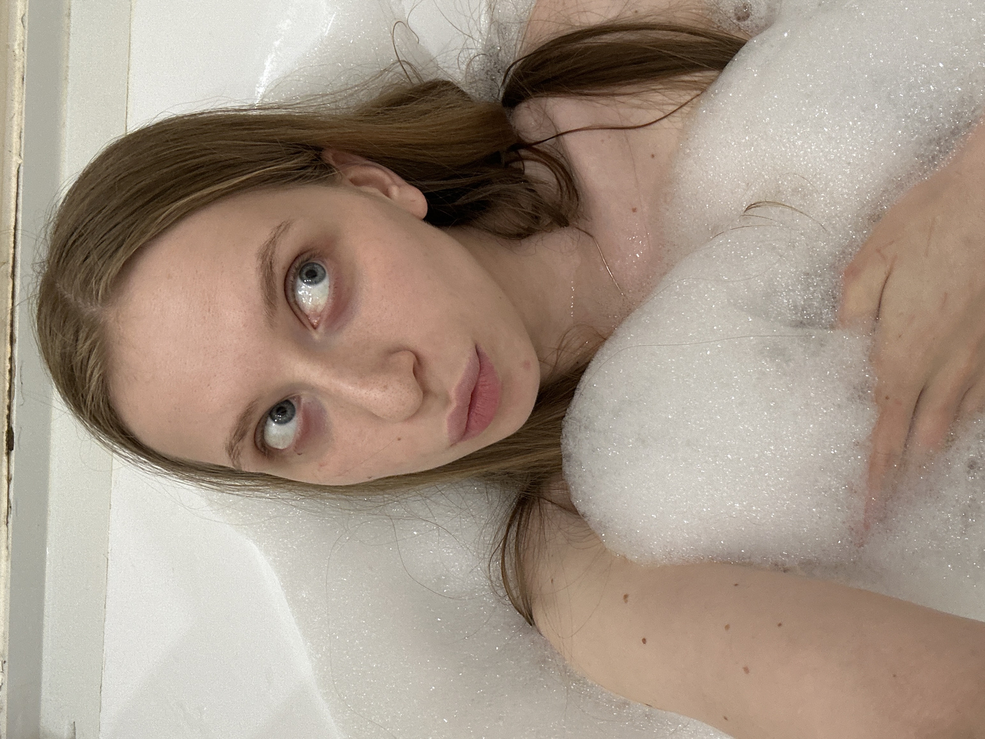A young, light-skinned woman with long brown hair is lying in a bathtub filled with white soap bubbles. Her head is positioned towards the left side of the frame, and her face is angled upwards. She has blue eyes and pink lips. Part of her body is covered by the bubbles. There is a glimpse of a white bathtub edge and wall on the left side of the image. The scene is indoors, presumably a bathroom. There is no indication the image was taken in Chaikovsky, Russia.