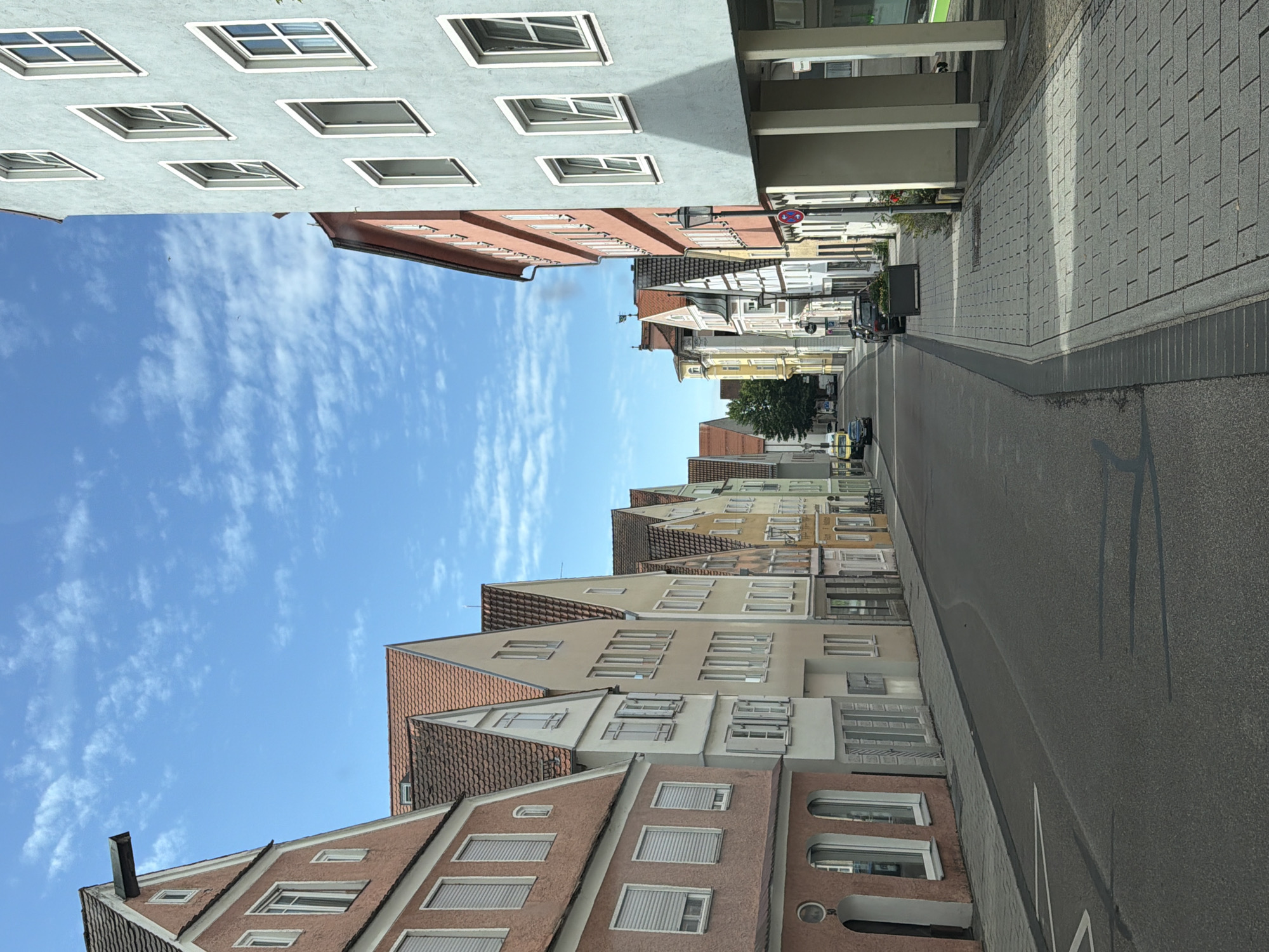 The image shows a street scene in Memmingen, Germany, with a row of colorful buildings on the left and a modern building with light blue facade on the right. The street is paved with asphalt and lined with a sidewalk made of grey paving stones. On the left side of the street, there are several old buildings with pitched roofs and various colored facades, predominantly in shades of pink, yellow, and cream. These buildings have traditional windows and some have shutters. On the right side of the street, a contemporary building with a light blue facade stands out. This building has multiple windows and is supported by large white pillars, creating a covered walkway. The sky is blue with scattered white clouds. Several cars are parked along the street and on the sidewalk. In the background, more buildings and trees are visible, suggesting a European town or city setting. The scene appears to be captured during daylight hours, under a clear sky.