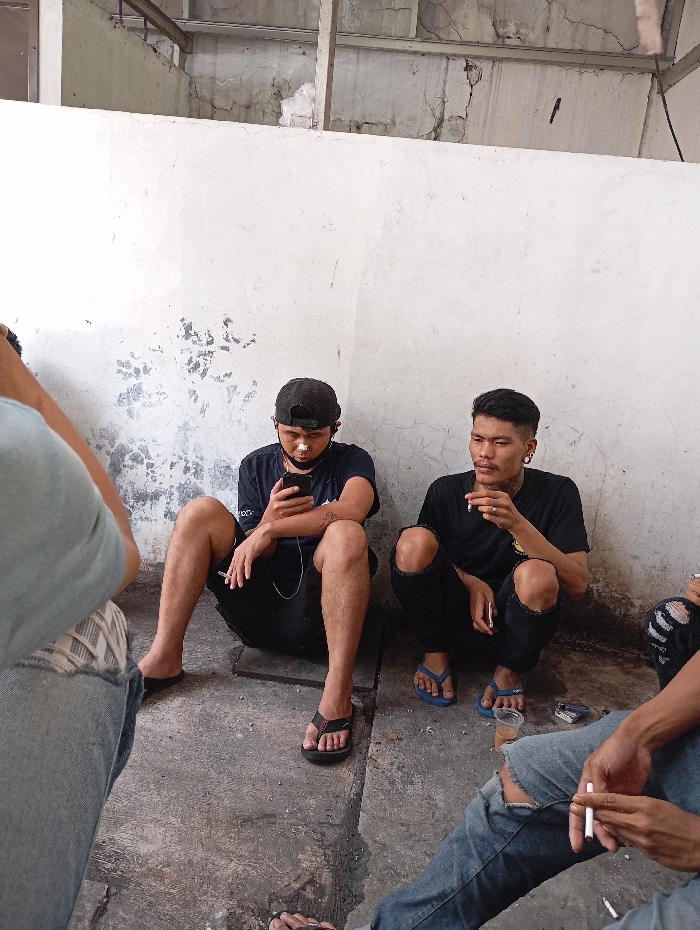 The image depicts a casual gathering of at least four men in what appears to be a utilitarian or informal concrete space in Katapang, Indonesia. Two men are prominently seated on the concrete floor, leaning against a weathered white wall with visible cracks and peeling paint.
The man on the left is wearing a black t-shirt, black shorts, a black baseball cap, and black flip-flops. He has a white patch or mask under his nose and is engrossed in his smartphone, which is connected to white wired earbuds. He holds a cigarette in his left hand.
Next to him, the man on the right is wearing a black t-shirt, ripped black pants, and blue flip-flops. He has short dark hair, ear gauges, and a light mustache. He is holding a lit cigarette to his mouth with his right hand and another unlit cigarette in his left. A small clear plastic cup containing a dark liquid and a cigarette pack are visible on the ground near him.
Partial views of other individuals are seen at the edges of the frame. On the far left, a person's shoulder, arm (in a light blue shirt), and legs (in light blue ripped jeans and sneakers) are visible. On the far right, legs in ripped blue jeans and an arm holding a lit cigarette are seen. The group appears to be socializing or taking a break. Most of the visible individuals are smoking cigarettes.
The setting includes a dirty concrete floor and an unfinished ceiling structure with beams and more cracked concrete above the wall. The lighting suggests it is daytime, casting a soft, ambient glow on the scene. The overall atmosphere is relaxed and informal. No discernible text is visible on clothing or surrounding objects.