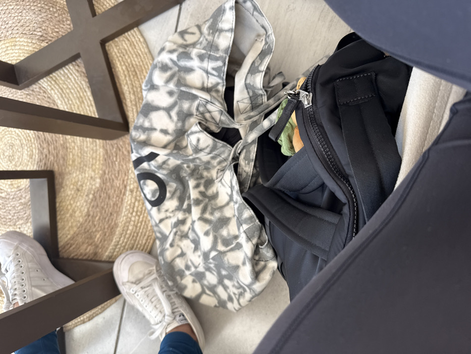 This low-angle, downward shot captures a person's lower body and belongings on the floor, likely taken from a seated position. The primary subjects are the person's legs and feet, clad in white sneakers. One leg, wearing dark leggings or athletic pants, is prominent on the right, while the other, wearing blue pants, is partially visible on the left.
Two bags rest on the light-tiled floor. On the left is a large, off-white tote bag featuring a grey, abstract, tie-dye or camouflage-like pattern. A large black "O" or "0" is distinctly visible on its side. To the right of the tote bag is a black backpack or duffel bag with a prominent silver zipper.
The setting appears to be an indoor environment, possibly a cafe or restaurant in Puebla City, Mexico. Dark brown, angular table legs are visible in the foreground, forming a geometric pattern. A circular, woven natural-fiber rug is partially visible beneath the table legs and feet, adding texture to the floor. The scene depicts a casual moment, with bags placed conveniently on the floor while the person is seated. The time of day, weather, and emotions are not discernible from the image.