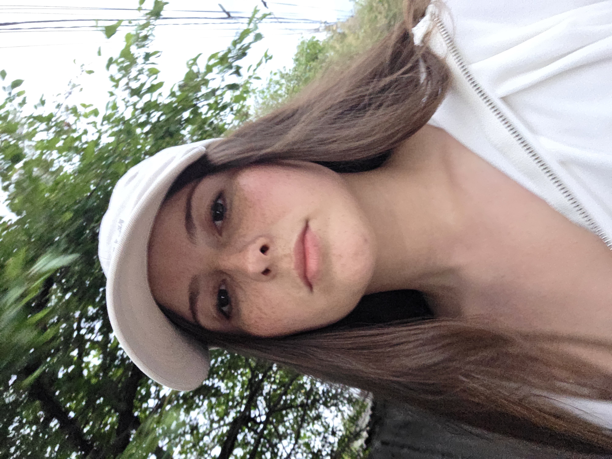 A young woman wearing a white baseball cap and a white zip-up hoodie poses for a selfie outdoors. She has long brown hair and is looking directly at the camera with a neutral expression. Freckles are visible across her nose and cheeks. The background consists of green foliage from trees and a slightly overcast sky, suggesting an outdoor setting in a non-urban environment.