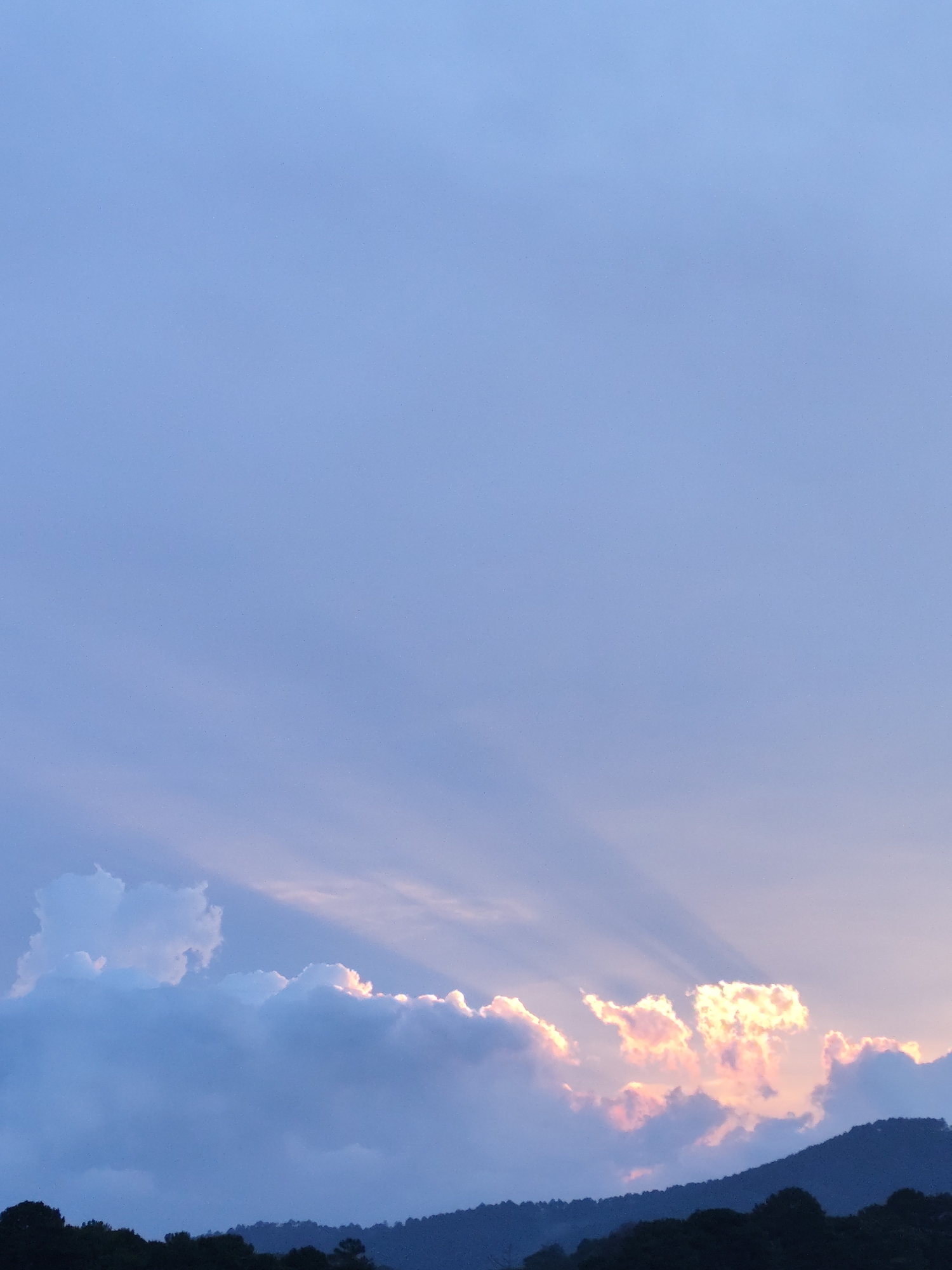 The image depicts a partially cloudy sky with a mountain range visible at the bottom. Sunlight streams through the clouds, creating crepuscular rays. The sky is predominantly blue, with a gradient towards lighter hues near the horizon. The mountain range is dark and silhouetted against the sky. The image was likely taken in Da Lat, Vietnam.