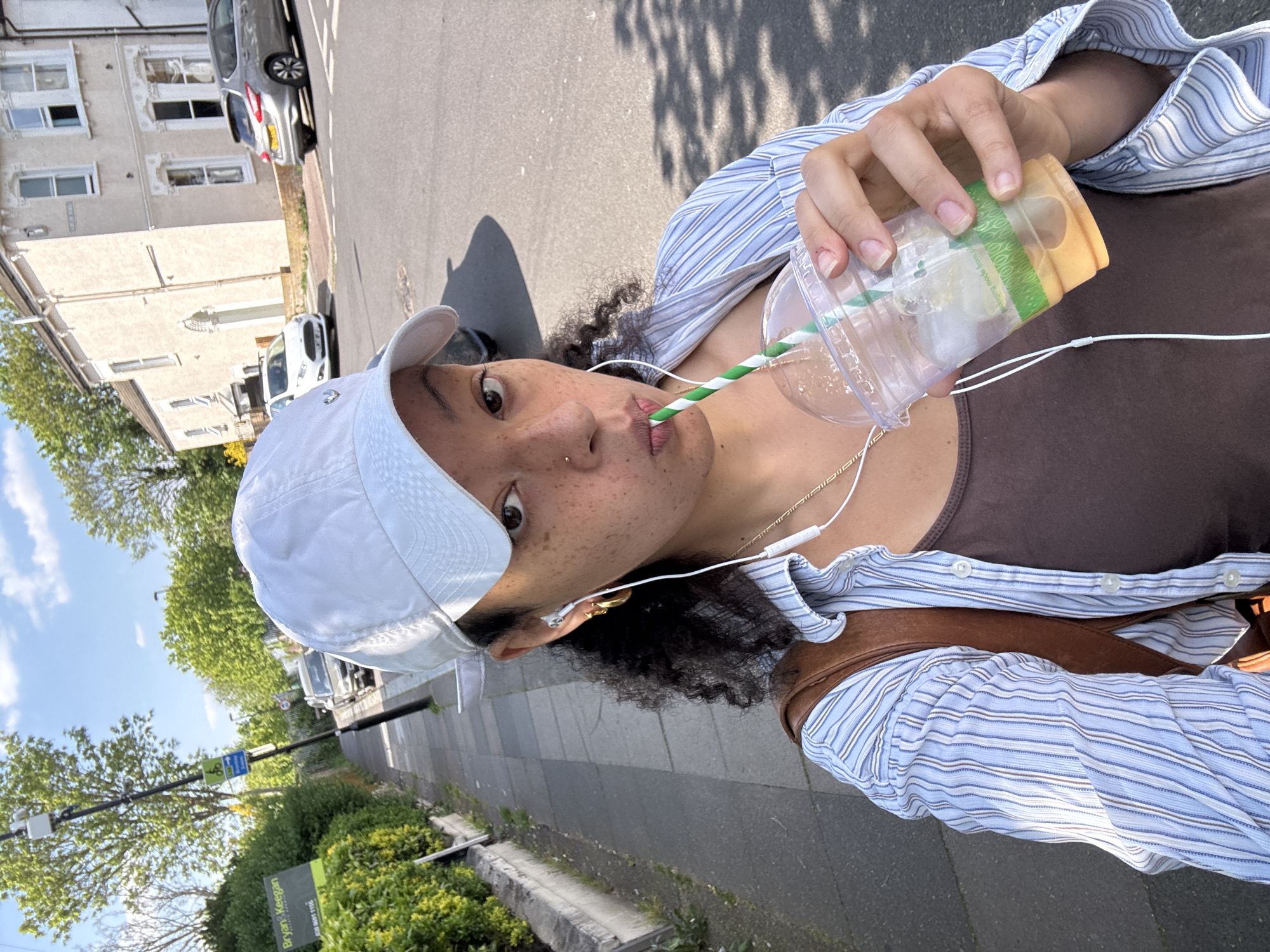 The image, taken in a selfie style and rotated clockwise, features a person outdoors on a sunny day in London, United Kingdom. The subject, appearing to be female, has dark, curly hair, freckles, and a nose piercing. She wears a white baseball cap, a light blue and white striped button-down shirt over a brown top, and a brown bag strap is visible across her shoulder. She holds a clear plastic cup filled with ice and a green and white striped straw to her mouth, appearing to drink from it. White wired earphones are also visible around her neck, and she wears gold earrings and a gold necklace.
The background reveals an urban street scene. On the left (when upright), residential buildings, possibly terraced houses with traditional window designs, line the street. Several cars are parked along the road, including a grey Ford Fiesta with a partially visible license plate ("YB13") and a white Ford Focus with the license plate "FM14 FORD FOCUS." The road itself has yellow double lines, indicating parking restrictions. On the right, a sidewalk or pavement is bordered by trees and bushes. A blue road sign displaying a bicycle symbol is present, alongside a green sign for "Bryans Keegan," an estate agent, which includes a phone number "020 8691 1155" and website "bryanskeegan.co.uk."
The bright sunlight and clear shadows on the ground indicate it is daytime, likely late morning or afternoon. The person looks directly at the camera with a neutral and candid expression. Text on the cup's band is partially visible, reading "making made from p," suggesting information about its material or origin.