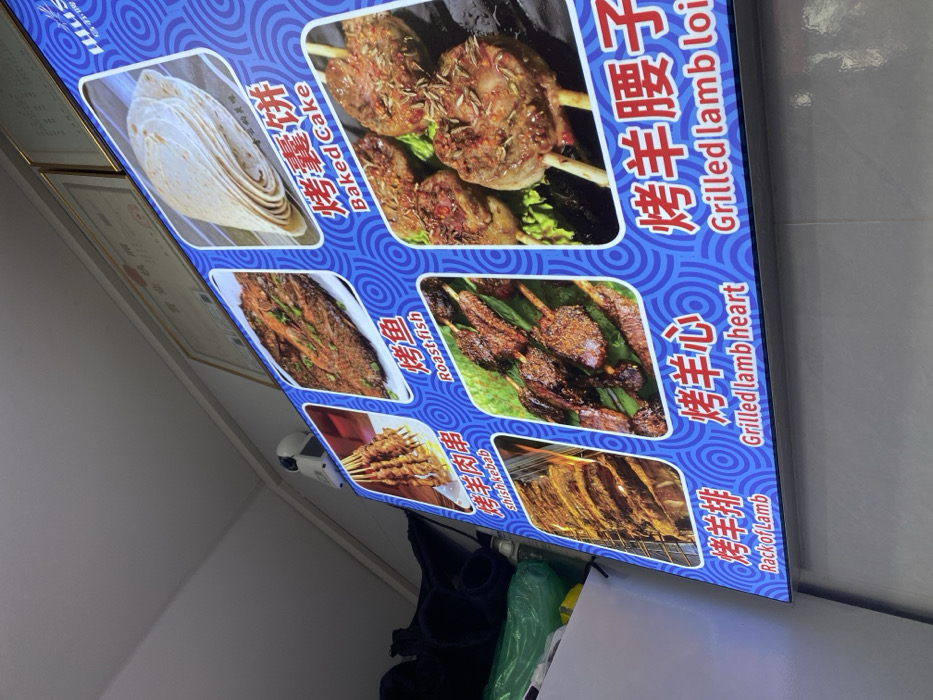 This media file displays a brightly lit, backlit menu board, likely from a restaurant or street food stall in Guang Zhou Shi, China. The board features a vibrant blue background with a circular pattern and showcases six different food items with accompanying photographs, Chinese names, and English translations.
The featured dishes include:
1. **烤囊饼 (Baked Cake):** Illustrated by a stack of round flatbreads.
2. **烤羊腰子 (Grilled Lamb Loin):** Pictured as chunks of grilled lamb on skewers, seasoned with herbs. The English translation is partially visible as "Grilled lamb loi".
3. **烤鱼 (Roast fish):** Shows a whole roasted fish, garnished with green herbs.
4. **烤羊心 (Grilled lamb heart):** Depicts skewers of grilled lamb heart, seasoned and charred.
5. **烤羊肉串 (Shish kebab):** Features multiple skewers of grilled meat, a common street food item.
6. **烤羊排 (Rack of Lamb):** Displays a rack of grilled lamb ribs, cooked to a golden hue.
The menu is designed for clear presentation, making it easy for customers to identify and choose dishes. A small white device, possibly a security camera or sensor, is visible below the menu board on the left side. The backlit nature of the display suggests it might be used in an indoor setting or during the evening/night.