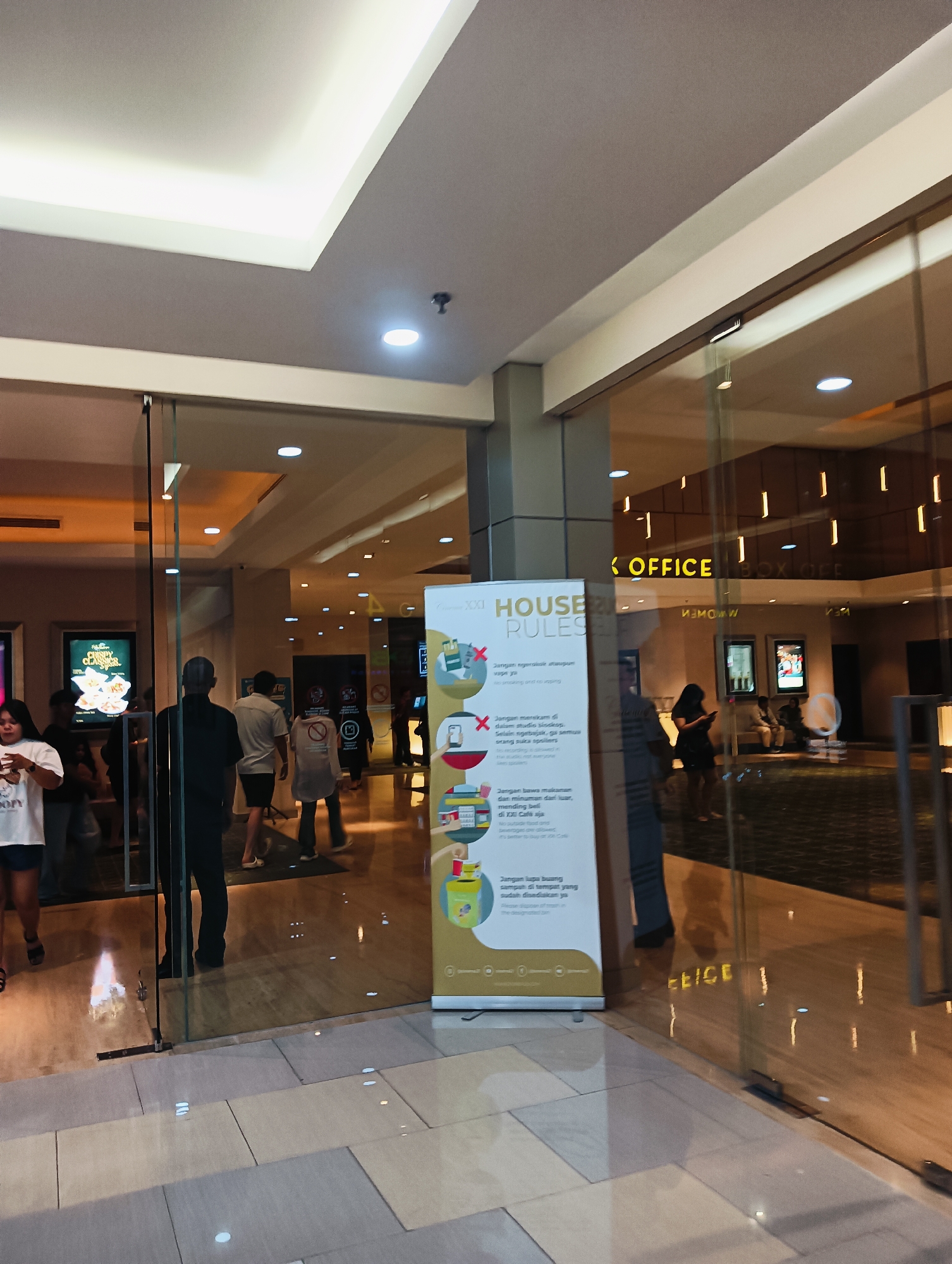 The image depicts the entrance area of a cinema complex, specifically XXI at Mega Mall Batam Centre, Indonesia. The scene is captured from a low angle, looking through a glass partition and door. Inside, several people are visible, some walking and others standing. A prominent display banner stands at the entrance, detailing "House Rules" in Indonesian, with illustrations of prohibited items like food, drinks, and phones.
The interior environment appears well-lit with recessed ceiling lights and decorative vertical strip lights. The flooring is a combination of polished wood-like material and lighter colored tiles, reflecting the overhead lights. Beyond the immediate entrance, the cinema lobby extends, with more people and signs visible, including what appears to be a "Box Office" sign reflected on the glass. The overall atmosphere suggests a public space within a shopping mall, likely during operating hours, with ongoing foot traffic.